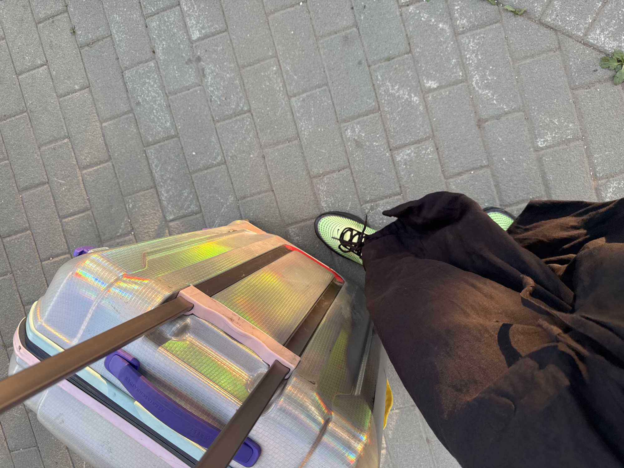 A top-down shot shows a person standing on a gray brick sidewalk next to a piece of luggage. The person is wearing black pants and neon green and black sneakers. The luggage is light gray with a clear, iridescent finish. It has a retractable handle with a light brown grip. The luggage also features purple and pink accents. The sidewalk is made of rectangular gray bricks, and some green weeds grow at the edge of the sidewalk in the upper right corner.