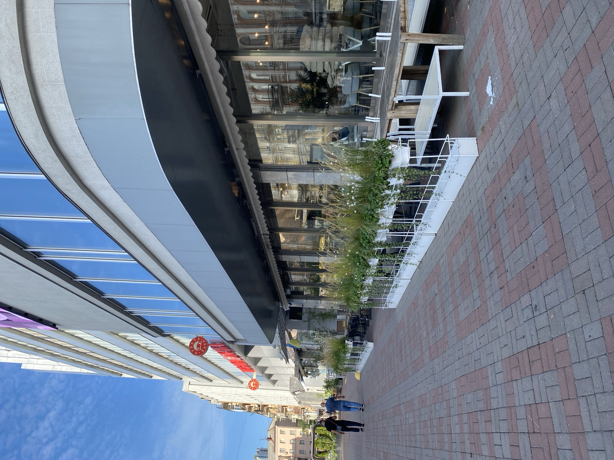 The image depicts a sidewalk scene in an urban environment, possibly a commercial district. The foreground is dominated by a red and gray brick-patterned walkway. To the right, a series of white planters filled with green foliage and tall grasses lines the sidewalk, creating a border. Behind the planters, a long wooden table with white benches is set up, suggesting an outdoor seating area for a cafe or restaurant. The building facade to the right features large glass windows that reflect the opposite side of the street, revealing architectural details of older buildings. An awning extends over the windows. Further down the sidewalk, two individuals are walking away from the camera. The building on the left is a modern, multi-story structure with numerous windows and a curved architectural element. Red circular signs with white text are visible on the building's facade. The sky is blue with some clouds.