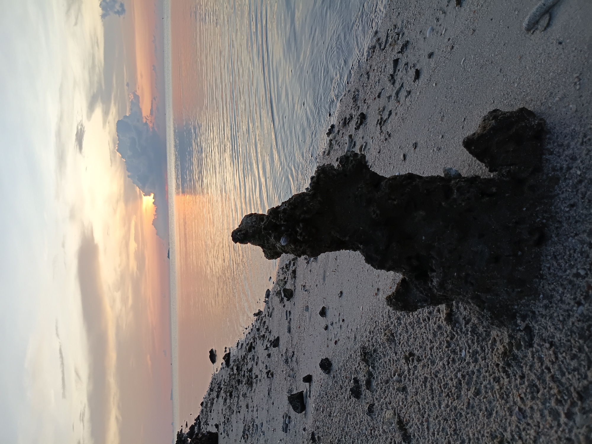The image depicts a sunset scene at Baan Wok Tum, Thailand. In the foreground, a dark, textured rock formation sits on a sandy beach. The beach is also scattered with smaller pebbles and debris. Beyond the beach, calm water reflects the colors of the sunset. The sky is filled with clouds, with the sun partially obscured behind one. The light from the sun creates a warm, orange glow that reflects on the water.