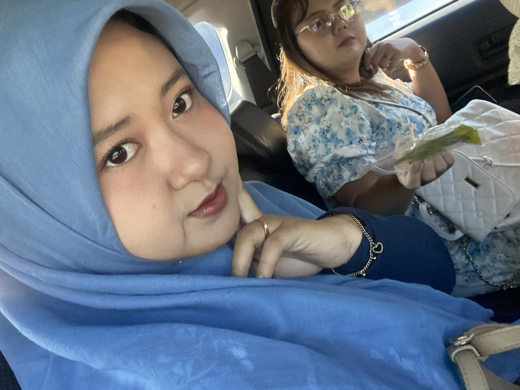 Two women are in a car. One in the front passenger seat is wearing a blue hijab and has a gold ring and bracelet. The woman in the back seat has glasses, a hair accessory, and is wearing a floral top. She is holding a white purse and something green. The location is possibly Muktisari, Indonesia.