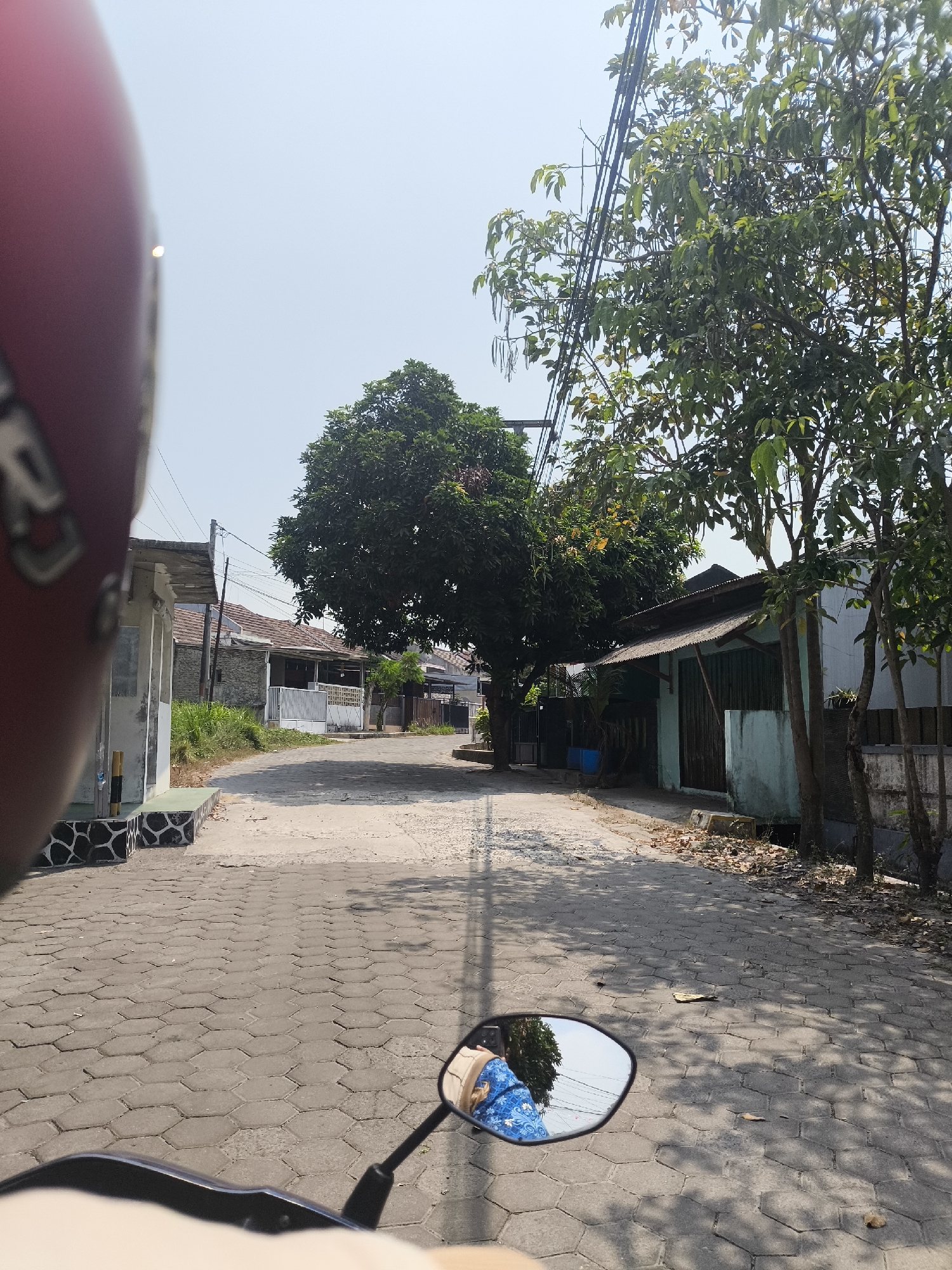 The image captures a view from a motorcycle moving along a paved street in a residential area, likely in Cilegon, Indonesia, on a clear, sunny day. The street is paved with hexagonal interlocking concrete blocks. On the left side of the road, there is a low building with a tiled roof and a patterned stone base. Further down the street, more houses with tiled roofs and fences are visible, interspersed with trees and utility poles. The right side of the street features trees with dense foliage and smaller buildings with corrugated metal roofs. A large, mature tree with dark green leaves dominates the center of the background. In the foreground, a portion of a red helmet is visible on the left, and a motorcycle's side mirror on the right reflects a person wearing a blue patterned garment, along with trees and the sky. Shadows from the trees are cast across the street, indicating the time of day. Debris, including dry leaves, is scattered on the sides of the road.