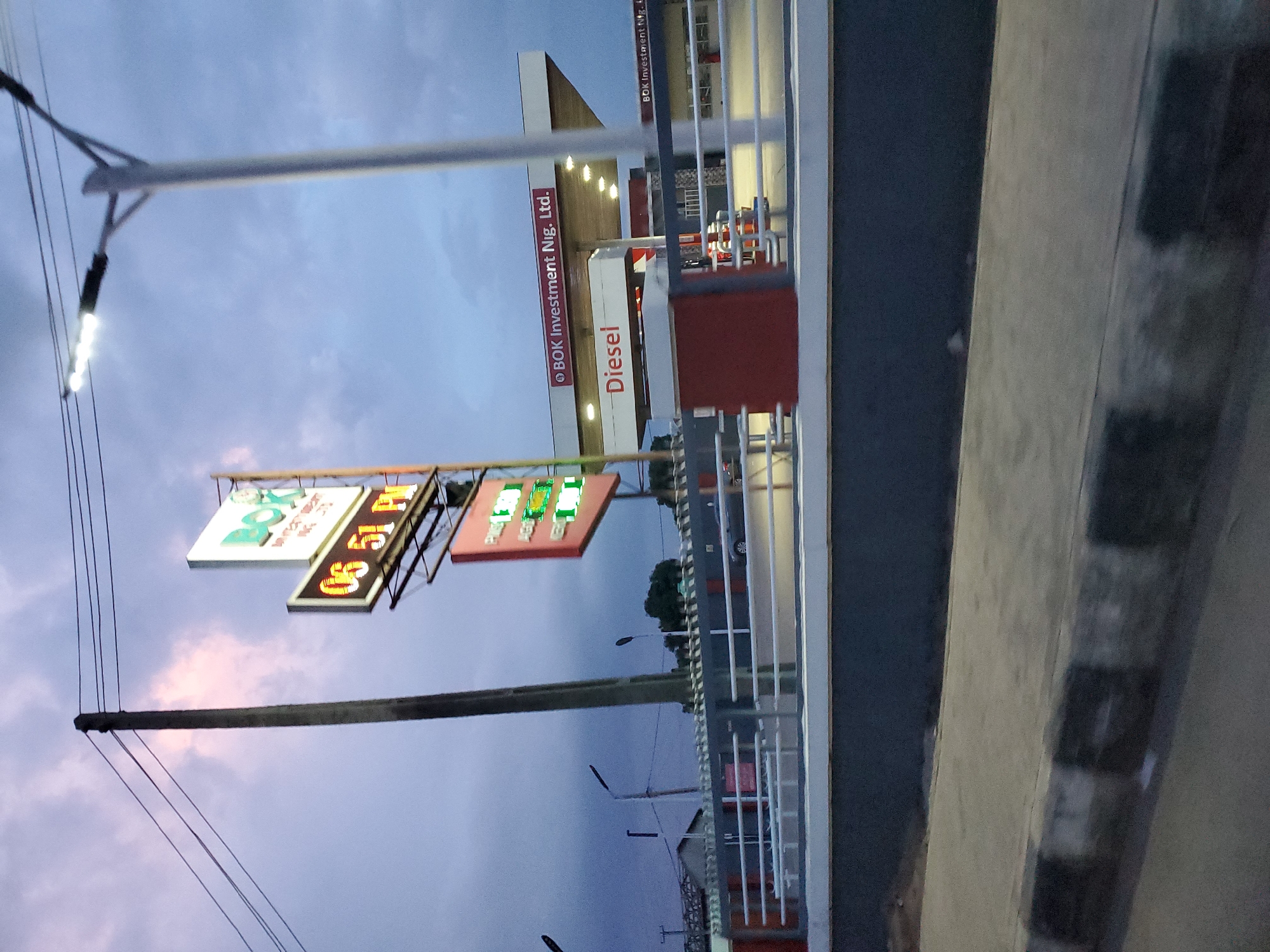 In Gura Top, Nigeria, a gas station's illuminated signage stands out against a twilight sky. A large sign displays "BOK Investment Nig. Ltd." and the time "06 51 PM." Below this, another sign indicates "Diesel." The gas station itself features a modern canopy with bright lighting. A street lamp with multiple bulbs is visible to the right. In the background, a fence with white railings separates the gas station from a road, where a dark-colored car is parked. The ground in the foreground is paved with concrete, featuring a curb with black and white stripes. The overall scene suggests a commercial area during the early evening hours.