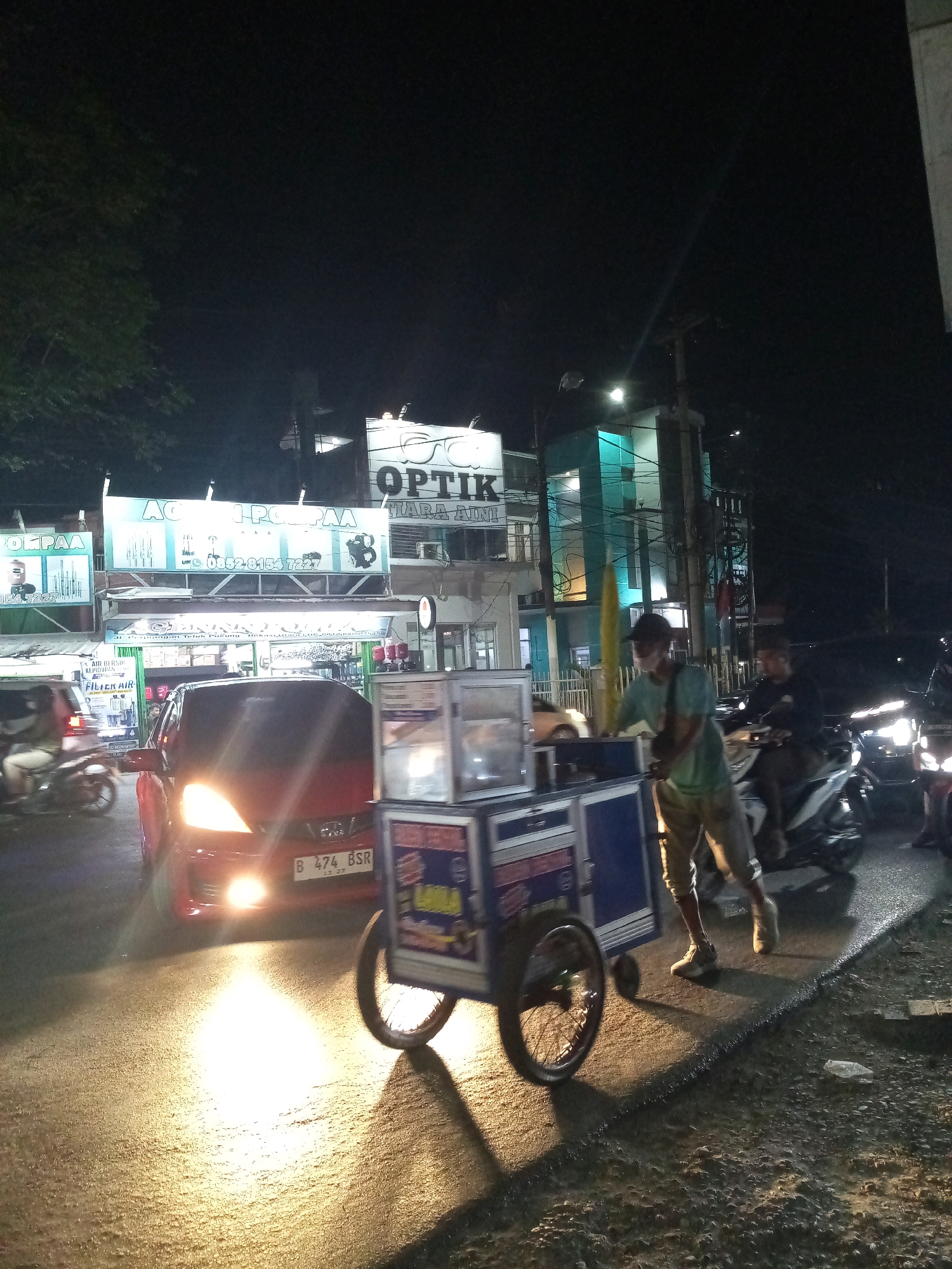 A nighttime street scene in Bekasi, Indonesia, depicts a busy commercial area with several businesses and vehicles. In the foreground, a person is pushing a food cart, appearing to be a street vendor. The cart has a blue and silver exterior with visible glass panels, suggesting it displays food items. The vendor is wearing a light green t-shirt, khaki shorts, and sneakers. Behind the vendor, a red car with its headlights on is stopped or moving slowly in traffic, its license plate clearly visible as "B 474 BSR". Several motorcycles are also present, some parked and others in motion, indicating a lively atmosphere.
Dominating the background are illuminated signs for various shops. One prominent sign reads "OPTİK TIARA AIRI" with an image of eyeglasses. Another sign, "AC.1 POMPAA" is also visible, suggesting sales of air conditioners and pumps. A teal-colored building stands behind the "OPTİK" sign, lit from within. The street itself is wet, reflecting the lights from the vehicles and businesses, indicating recent rain or a damp surface. The overall impression is of a typical urban streetscape at night, with commercial activity and pedestrian/vehicle traffic.
