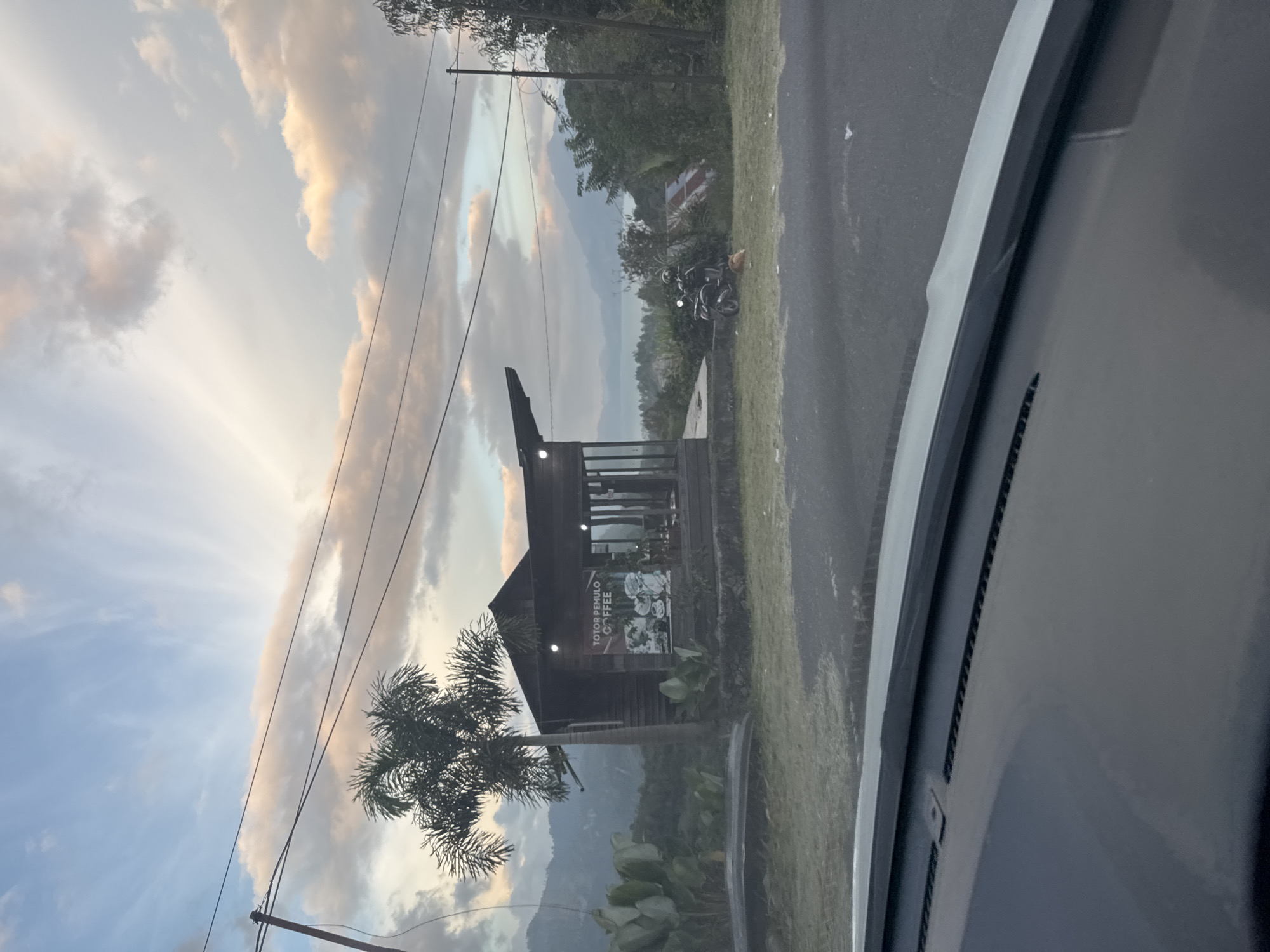 The image depicts a scenic view from a vehicle's dashboard, looking out at a small wooden cafe situated on a hillside. The cafe, labeled "TOTOR PEMULO COFFEE," has large windows and an overhang. A palm tree stands to the left of the cafe, with lush green foliage and distant hills in the background. The sky is a mix of blue and clouds, with hints of sunset colors. A motorcycle is parked near the cafe, and a cat is resting on the grass. The foreground shows the interior of a car, with its dashboard visible. The location is specified as Payatumpi Baru, Indonesia.