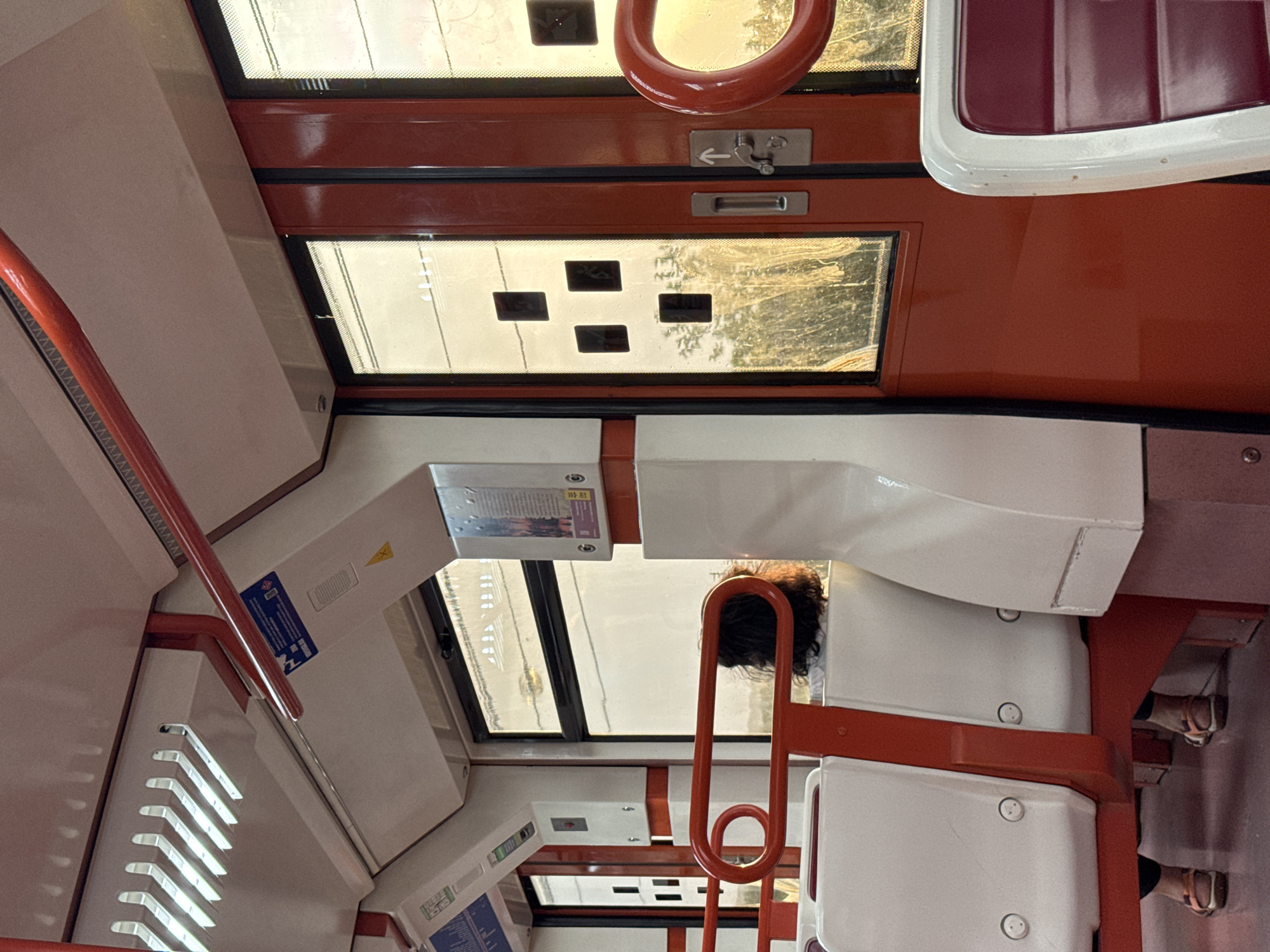 The interior of a train carriage on the Madrid Metro, likely in Arganda del Rey, Spain, is depicted. The carriage features white and red seating, red handrails, and large windows. Sunlight streams in through the windows, creating a warm, golden hue and casting reflections on the glass. A person with dark hair is seated facing away from the camera, looking out the window. The windows have a grid pattern of dark squares superimposed on them, possibly indicating informational displays or decorative elements. The overall impression is one of a peaceful commute during sunset.