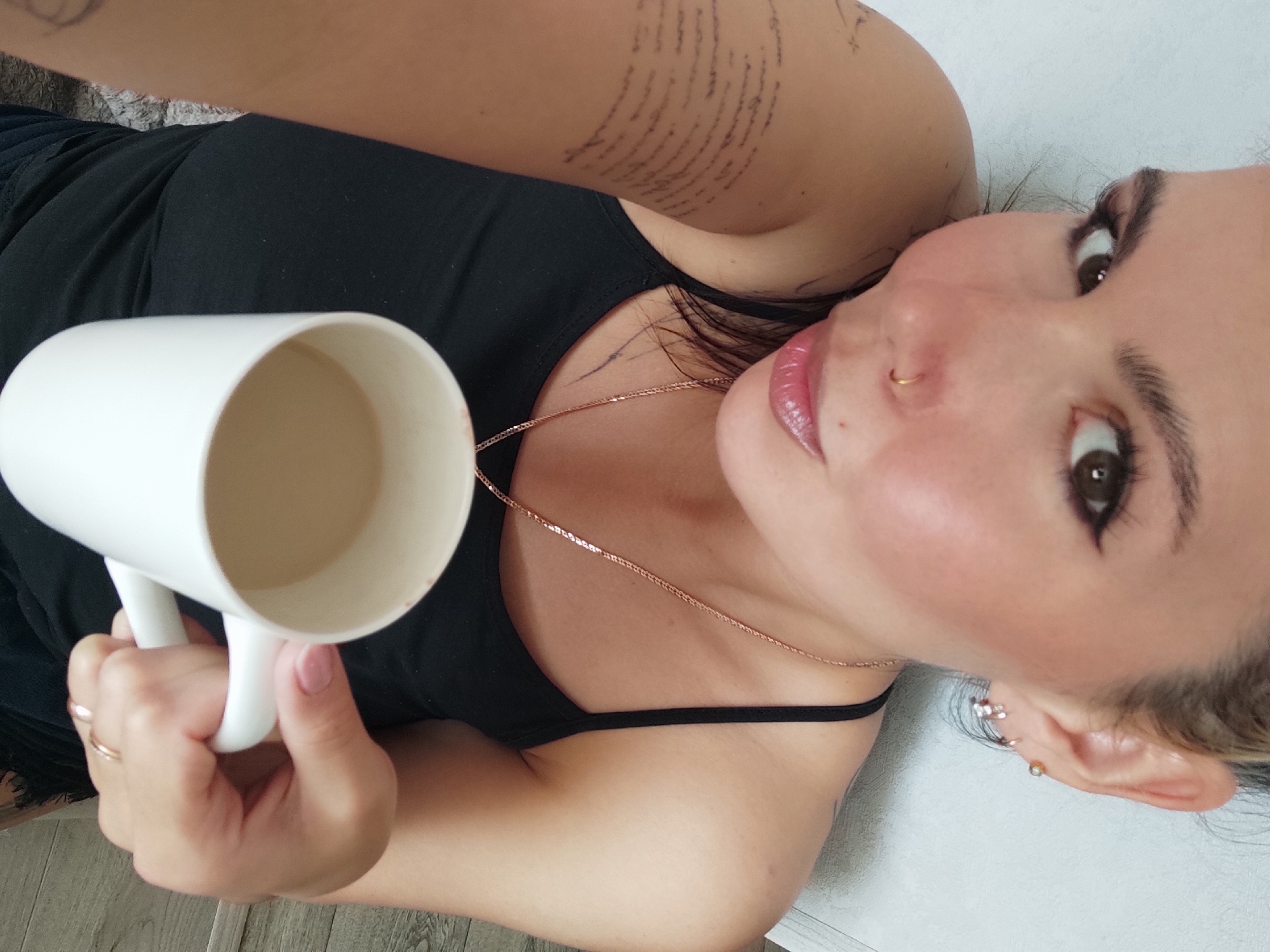 A woman is lying on a light-colored surface, possibly a bed or floor, holding a large white mug in her right hand. She is wearing a black tank top and a delicate gold chain around her neck. Her left arm is visible, with text tattooed on the inner arm. She has pierced ears with multiple earrings and a nose piercing. The woman is looking at the camera with dark eye makeup. The environment appears to be indoors, with a glimpse of a wooden floor in the lower left corner. The city of Chaikovsky, Russia, is not discernible from the visual data provided.