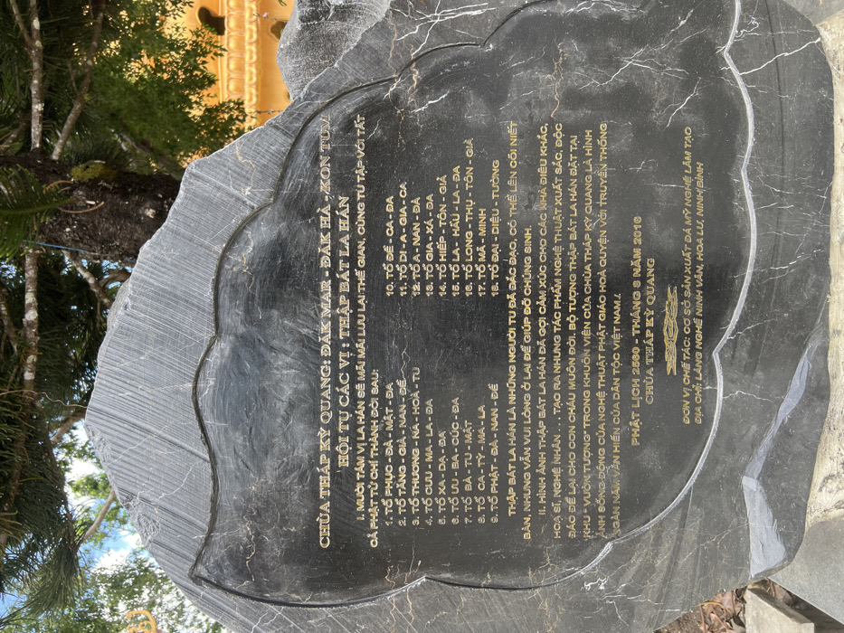 A large, irregularly shaped dark grey marble or granite stone tablet, with a polished surface, stands outdoors at Kỳ Quang Pagoda in Đăk Mar, Đăk Hà District, Kon Tum Province, Vietnam. Golden Vietnamese text is inscribed on its face, identifying the location and title "HỘI TỤ CÁC VỊ: THẬP BÁT LA HÁN" (Gathering of the Eighteen Arhats). A list of 18 names is presented in two columns. Additional text describes the significance and creation of the Arhat sculptures, dated Buddhist Calendar 2560 (August 2016), and credits the stone art production facility in Ninh Vân, Hoa Lư, Ninh Bình. The stone is positioned in front of a large tree with visible branches and green foliage. To the right, a golden, ornate architectural detail, likely part of a pagoda building, is partially visible. No individuals are present in the scene.