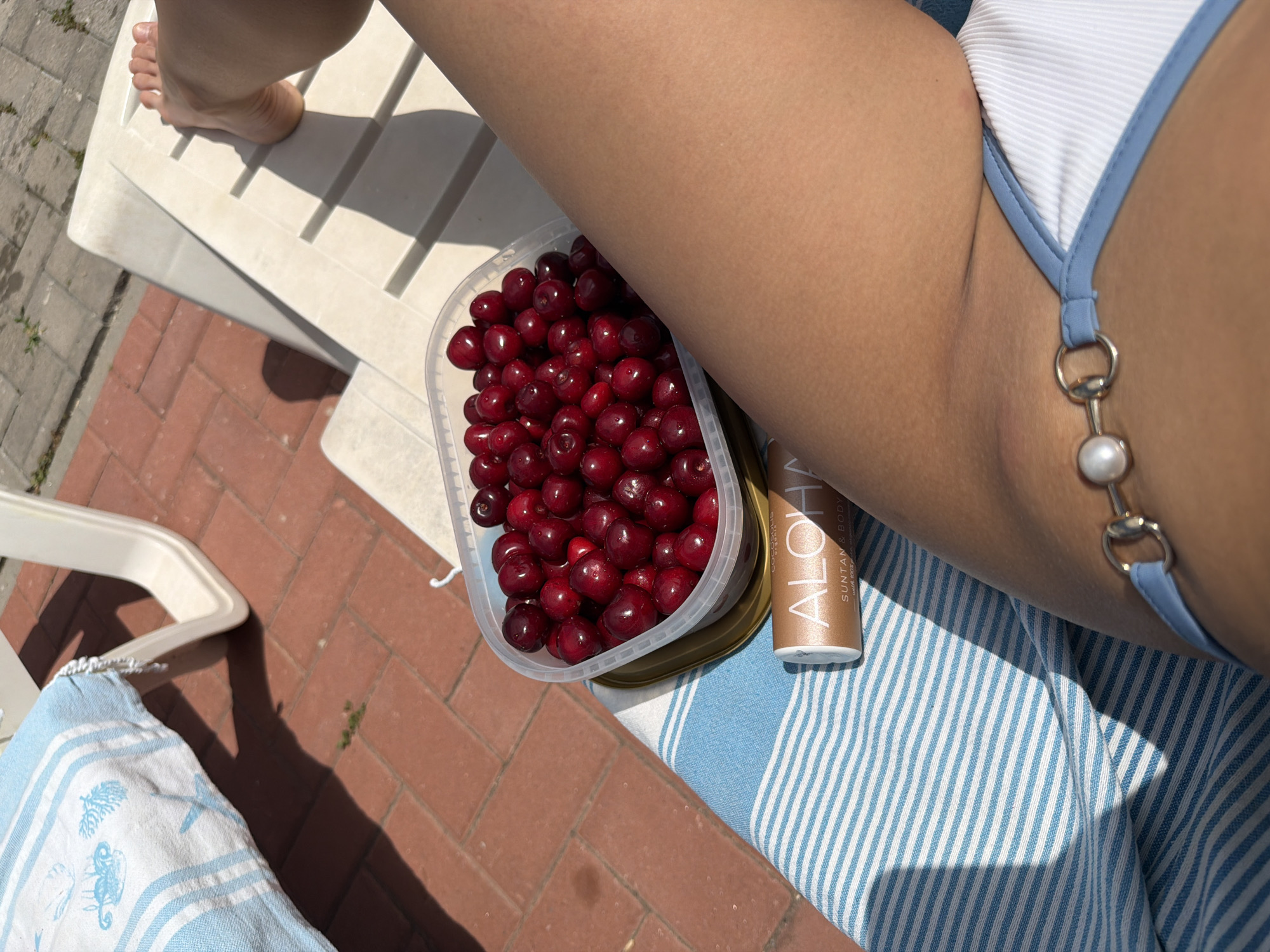 A person's tanned leg, wearing a white bikini bottom with a pearl and chain detail, rests on a white plastic lounge chair on a sun-drenched patio with red brick paving stones. A blue and white striped towel is draped over the chair. To the left of the leg is a clear plastic container filled with fresh cherries. Next to the cherries is a tube of "ALOHA" suntan and body oil. In the background, the corner of another lounge chair with a blue and white striped cushion is visible. The scene suggests a relaxed outdoor setting, likely in Simitli, Bulgaria, on a sunny day.