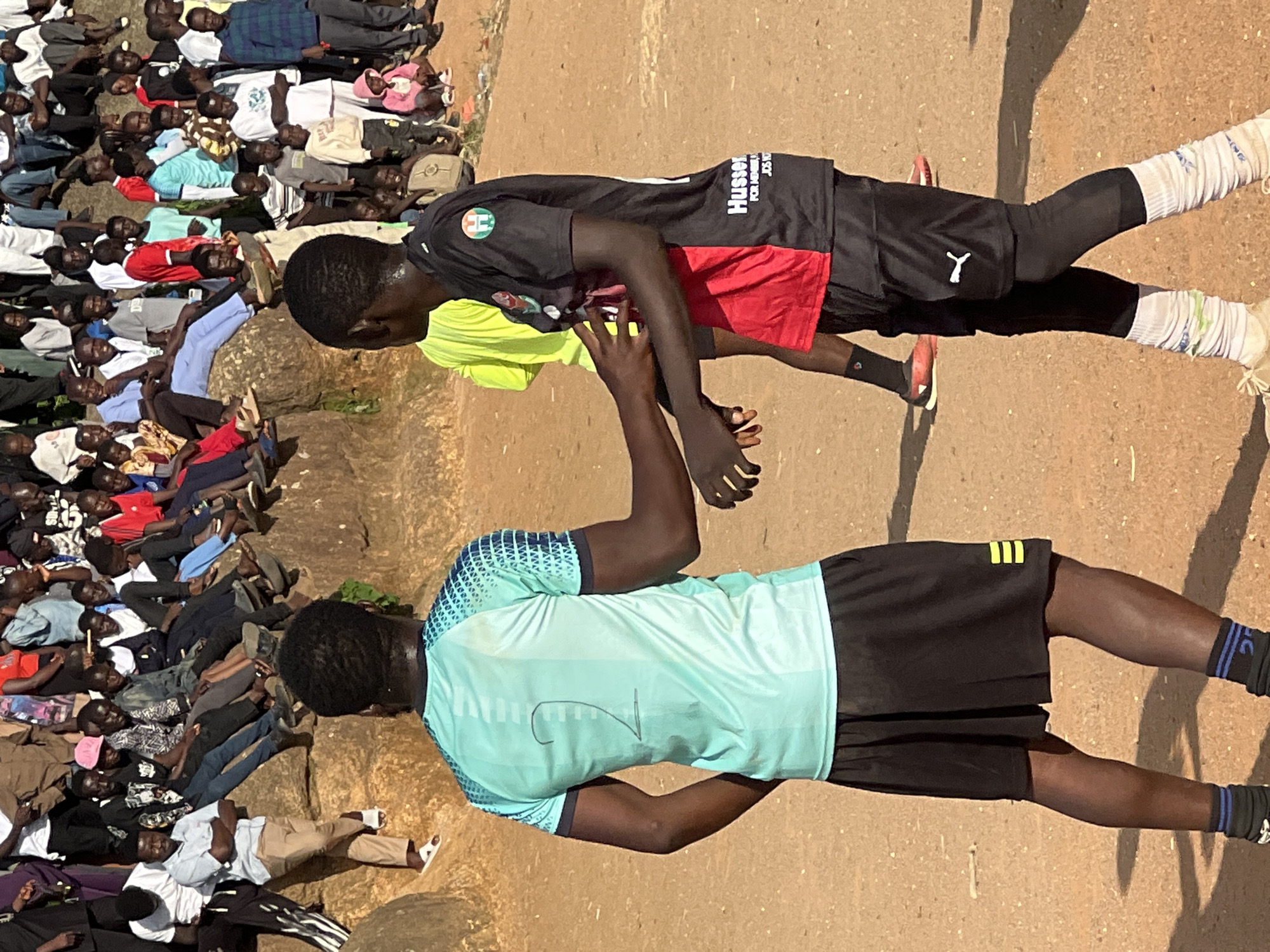 In Jos, Nigeria, two young men are the focus of this outdoor scene. The man on the left, facing away from the camera, wears a light blue t-shirt with a number "2" drawn on the back and black shorts. The man on the right, partially visible and facing left, is wearing a black and red soccer jersey and black shorts with white socks and cleats. They appear to be interacting, possibly during a game or practice. A crowd of people, mostly young men and boys, are gathered behind them, seated and standing on a rocky incline, suggesting a local sporting event or gathering. The environment is earthy and appears to be a dusty outdoor space with some rocks and sparse vegetation.