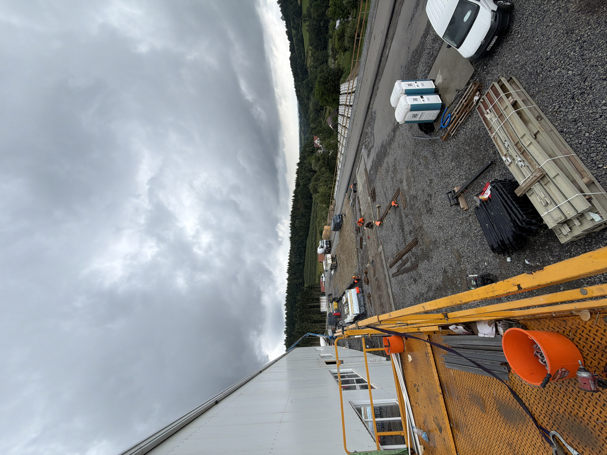 This is a wide-angle, high-angle, exterior, daytime shot of a construction site in Stróża, Poland. The scene is dominated by a large, white industrial building on the left, extending out of frame. In the foreground, a yellow construction platform or scaffolding is visible, with an orange bucket filled with chains, a power tool, and various construction materials. The ground is covered in gravel, with stacks of rebar and other building components scattered throughout. Several portable toilets are set up near a paved area. In the mid-ground and background, there are various construction vehicles, including a white van, a cement truck, and other machinery. A dense, green forest lines the horizon, with some houses visible in the distance amidst rolling hills. The sky is overcast with heavy, dark clouds, suggesting a cloudy or rainy day. There are several construction workers visible in the mid-ground, wearing orange safety vests and actively working on the site, some appearing to be laying rebar. The overall impression is one of an active construction project in a semi-rural, hilly landscape.