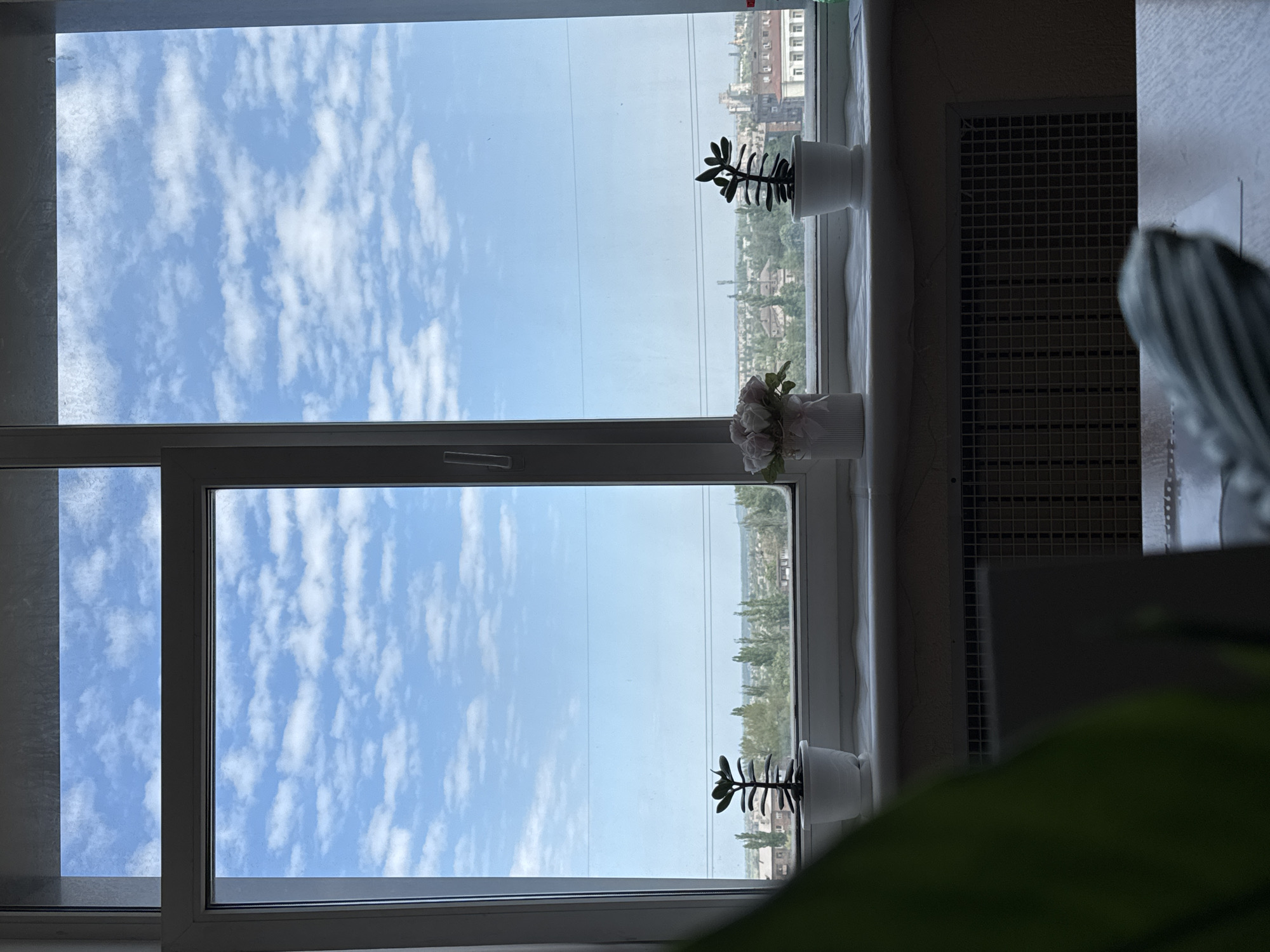 A view from inside a room, looking out through a window with a white frame divided into two panes. The sky is visible through the window, with scattered white clouds against a blue background. On the windowsill, there are three small white pots containing plants: two with succulent-like foliage and one with artificial roses. In the background, beyond the plants and the sky, is a cityscape with buildings and trees. The overall location appears to be Kryvyi Rih, Ukraine, judging by the style of buildings and general environment. A portion of a dark-colored radiator and a white wall can be seen to the right of the window, partially obscured by a large green plant. The photograph has a bright, airy feel.