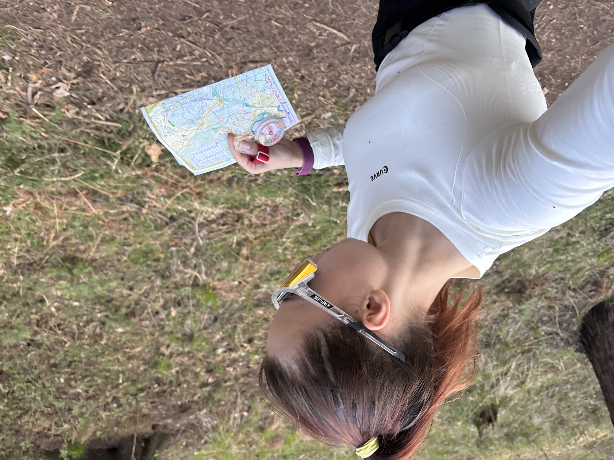 A woman is standing in an outdoor location with a map and compass in her hand. The map shows topographical features, and the compass is red and silver. The woman is wearing a white long-sleeved shirt with the word "CURVE" on the left side of her chest, sunglasses with yellow lenses, and her hair is pulled back with a yellow hair tie. She appears to be using the map and compass for navigation. The ground is covered with grass, dry leaves, and small branches, suggesting the location is in a wooded or park area. The image was taken near the city of Perm, Russia.