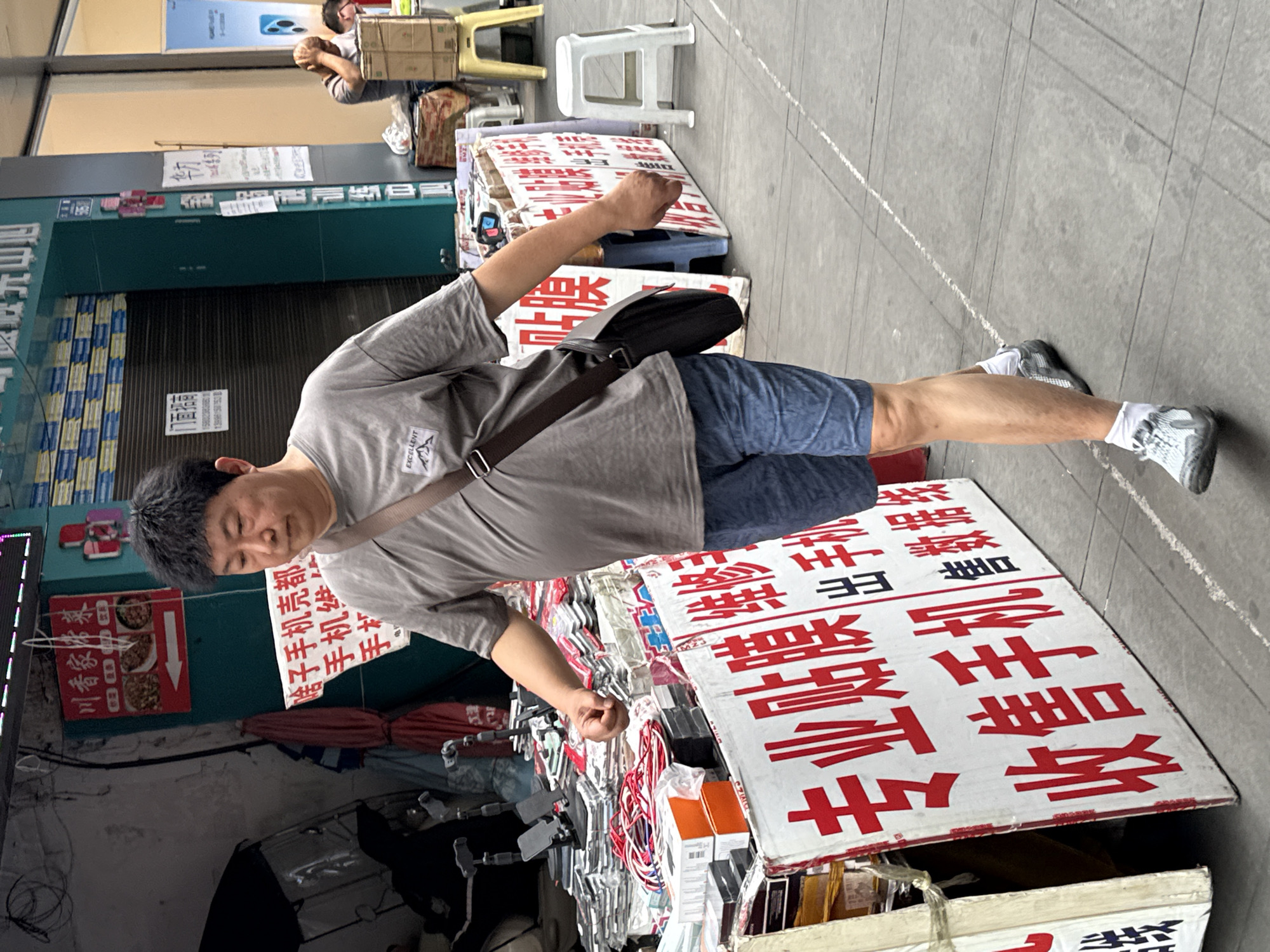 An eye-level, full outdoor shot of a man standing on a sidewalk in Jiangjin, China. The man has short, dark hair and is wearing a grey t-shirt, blue shorts, and grey and white sneakers. A black bag is slung across his body. He appears to be standing in front of a table laden with goods for sale. On the table are boxes, phone cases, and signs in Chinese characters. Behind the man, a store with closed shutters can be seen. A sign with a menu is visible. Further back, there is another person sitting down. A white plastic stool is on the sidewalk. The environment is urban, with a grey sidewalk and buildings in the background.