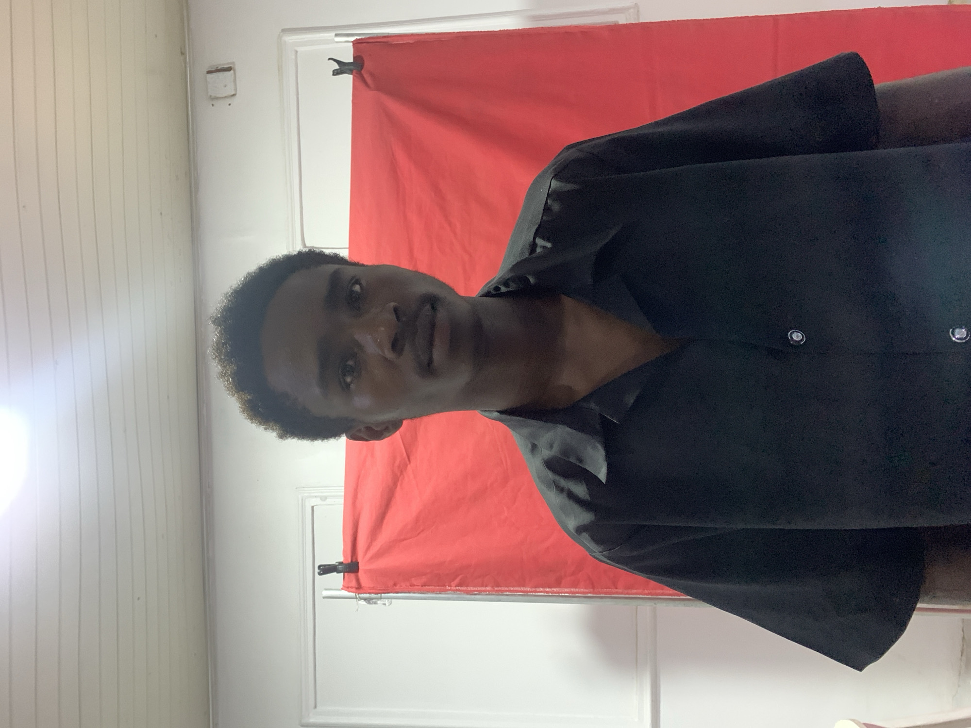 A young man with dark skin and short, dark, curly hair is wearing a black button-down shirt. He is standing in front of a red backdrop held up by a white rod. The background wall is white and appears to be made of wood paneling. A white doorframe and wall are visible to the right. This is likely a makeshift photography setup indoors. The location is unknown, so I cannot confirm that this is in Jos, Nigeria.