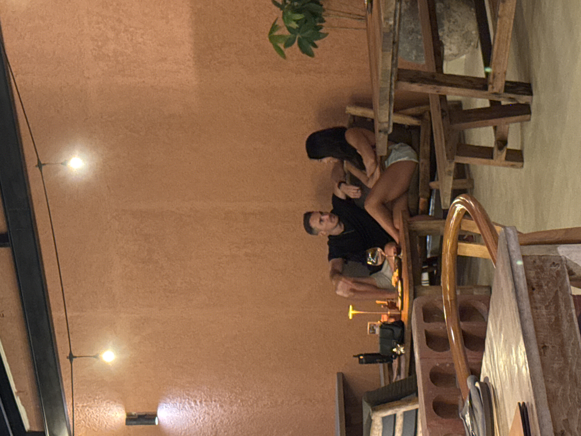A man and woman are seated at a table in an indoor space with tan-colored walls, likely a restaurant. The man wears a black shirt and is holding a glass of wine, while the woman, who has dark hair, is wearing a white top and a short bottom. The seating appears to be wooden and of a rustic style, with multiple chairs stacked nearby. The environment is illuminated by light fixtures, and there is a plant visible behind them. The location is Ungasan, Indonesia.