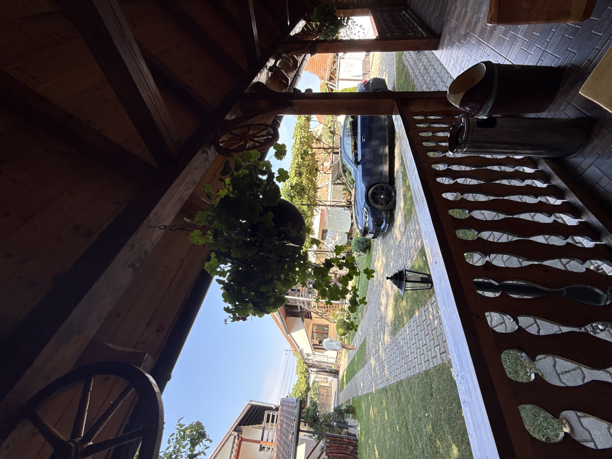 The image depicts a residential area in Cârțișoara, Romania, on a clear, sunny day. In the foreground, a wooden porch with a decorative railing is visible, furnished with two trash receptacles. Beyond the porch, a paved driveway leads through a grassy yard lined with houses. A dark blue sedan is parked on the driveway. A person dressed in light-colored clothing is walking away from the camera down the driveway. The scene is framed by wooden beams and hanging greenery, with a large wagon wheel decorative element visible on the porch ceiling.