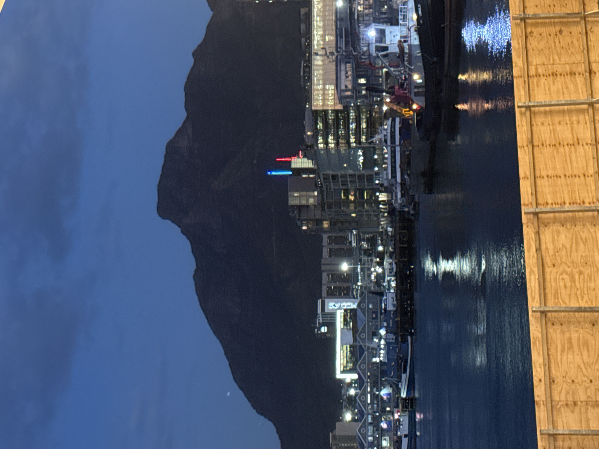 The image captures a vibrant night scene of a city's waterfront, with a majestic mountain silhouetted against a dark blue twilight sky. The perspective is likely taken from across the water, looking towards the illuminated city and the mountain.
The central subject is a dramatic, dark mountain range, characteristic of Cape Town's topography (likely Table Mountain or a contiguous peak), which looms large on the left side of the frame. Its rugged outline is visible against the deep blue, cloudless sky, suggesting clear weather conditions.
Below the mountain, a strip of modern city buildings lines the waterfront. These buildings are brightly lit with numerous artificial lights, casting warm yellow, white, red, and blue glows. Reflections of these city lights shimmer and stretch across the calm, dark waters of what appears to be a harbor or bay. On one of the taller buildings, prominent red and bright blue vertical lights stand out, adding a distinct focal point to the urban landscape. Faintly visible on a building sign, the letters "MEW" can be discerned, and below that, what appears to be "V&A" is suggested, strongly pointing to the V&A Waterfront in Cape Town.
In the immediate foreground, occupying the right side of the image, is a tall, light-colored wooden structure, possibly temporary scaffolding or a construction barrier, creating a stark contrast with the illuminated city beyond.
The overall scene conveys the bustling atmosphere of an urban center during the evening or night, set against the impressive natural backdrop of Cape Town, South Africa. The artificial lights create a sense of activity and warmth, mirrored by the tranquil water.