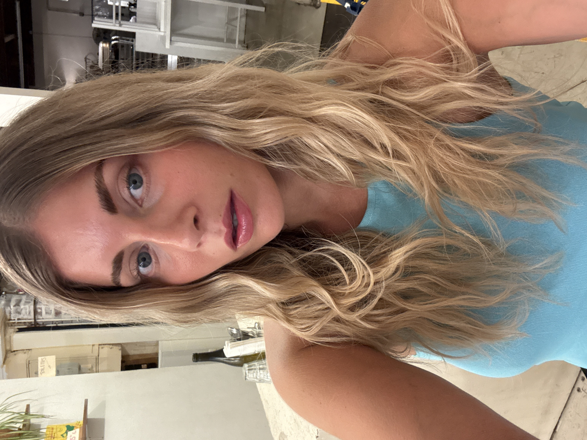 A close-up, waist-up, outdoor, selfie-style photograph shows a young woman with long, wavy blonde hair. She is wearing a light blue, ribbed tank top and is looking towards the camera with a neutral expression. Her makeup includes defined eyebrows, visible eyeliner, and glossy pink lipstick. Her right arm is visible on the left side of the frame, resting on a table. Behind her, a blurry background suggests an indoor setting, possibly a restaurant or cafe, with shelves containing bottles and glasses. A sign with French text is partially visible on a wall in the upper left quadrant. The lighting appears to be natural and diffused. The woman's skin is lightly tanned.