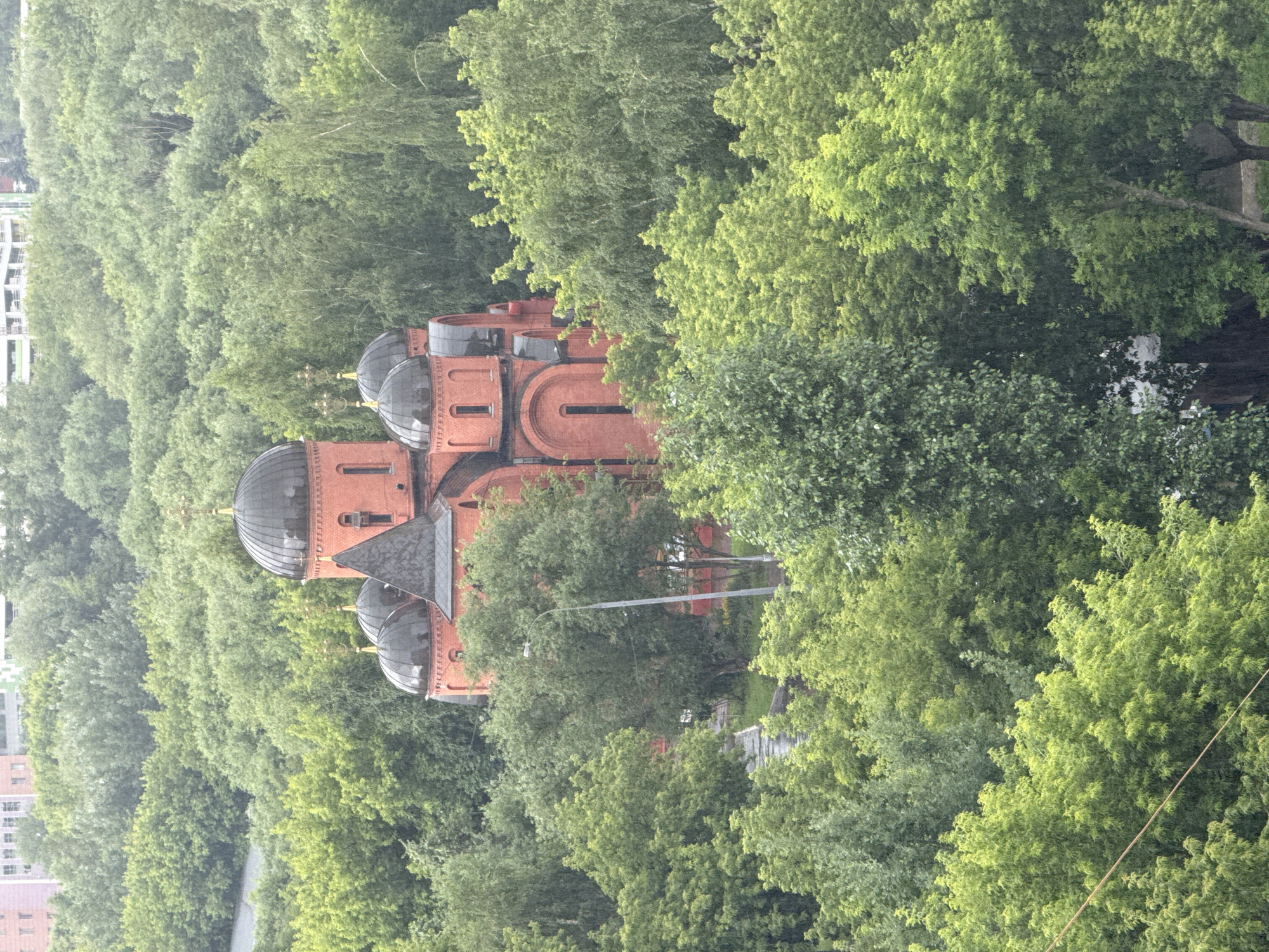 This is a vertical, high-angle shot of an ornate, red brick building, likely a church or cathedral, nestled amongst lush green trees. The building features multiple domes, some dark and rounded, and others smaller and topped with crosses. The architecture suggests a historical or religious site. The dense foliage of the surrounding trees creates a natural frame for the building, obscuring parts of it and giving the impression of a secluded or well-integrated structure within a park or forested area. The lighting is diffused, suggesting an overcast day, possibly in the late morning or afternoon. There are no visible people or explicit activity. The overall mood is one of tranquility and historical presence, enhanced by the natural setting. The location is identified as Moscow, Russia.