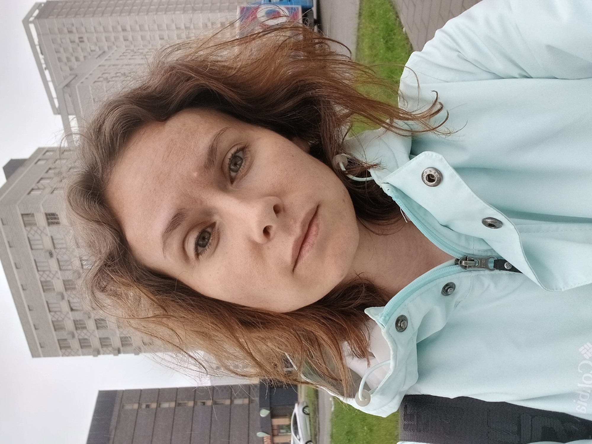 A selfie taken by a woman with brown, wavy hair, looking at the camera with a neutral expression. She is wearing a light blue jacket with a zipper and buttons. In the background, there are several tall buildings and a street with a sidewalk and grass. The sky is overcast, suggesting a cloudy day. The image appears to be taken outdoors in an urban environment. Based on the architecture and the presence of the "Columbia" logo on her jacket, the location could be Yekaterinburg, Russia, which is known for its modern architecture and some Columbia sportswear retailers.