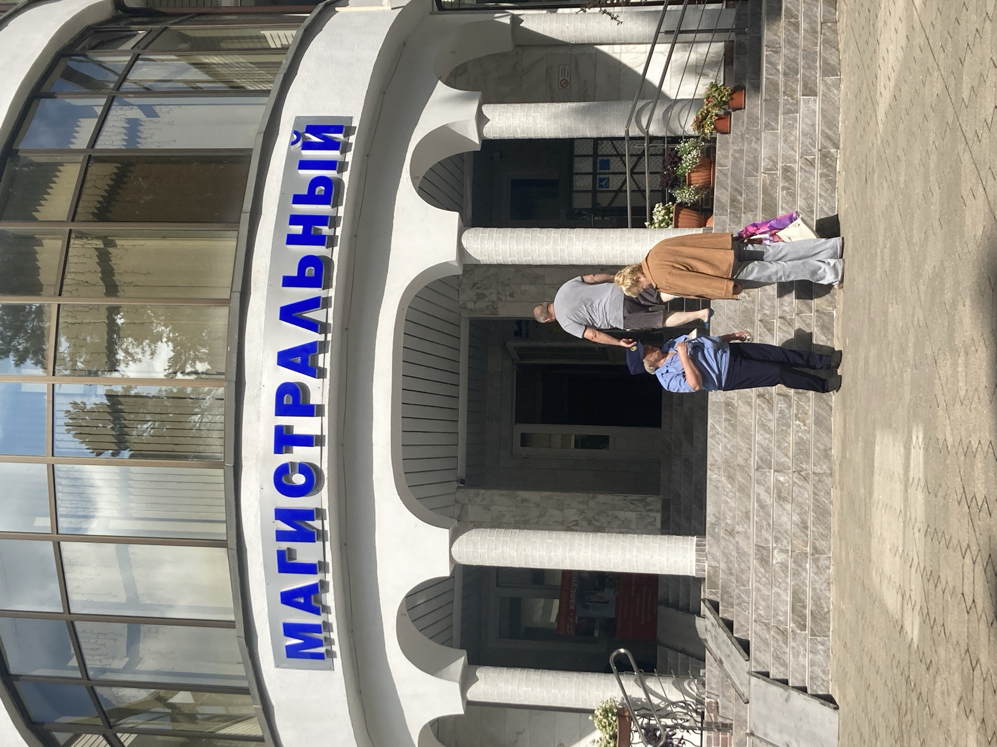The image shows the exterior of a building with blue lettering that reads "МАГИСТРАЛЬНЫЙ" (Magistralny). The building has a modern facade with large windows and arched entrances supported by white columns. Several people are visible on the steps leading up to the building. On the left, a woman in a blue uniform, possibly a police officer, stands facing a woman in a brown jacket and gray pants carrying a bag. Behind them, a man in a gray t-shirt and dark shorts is walking up the stairs. The scene appears to be a sunny day. The building is located at vulica Brestskaya 108, Baranavichy, Belarus.