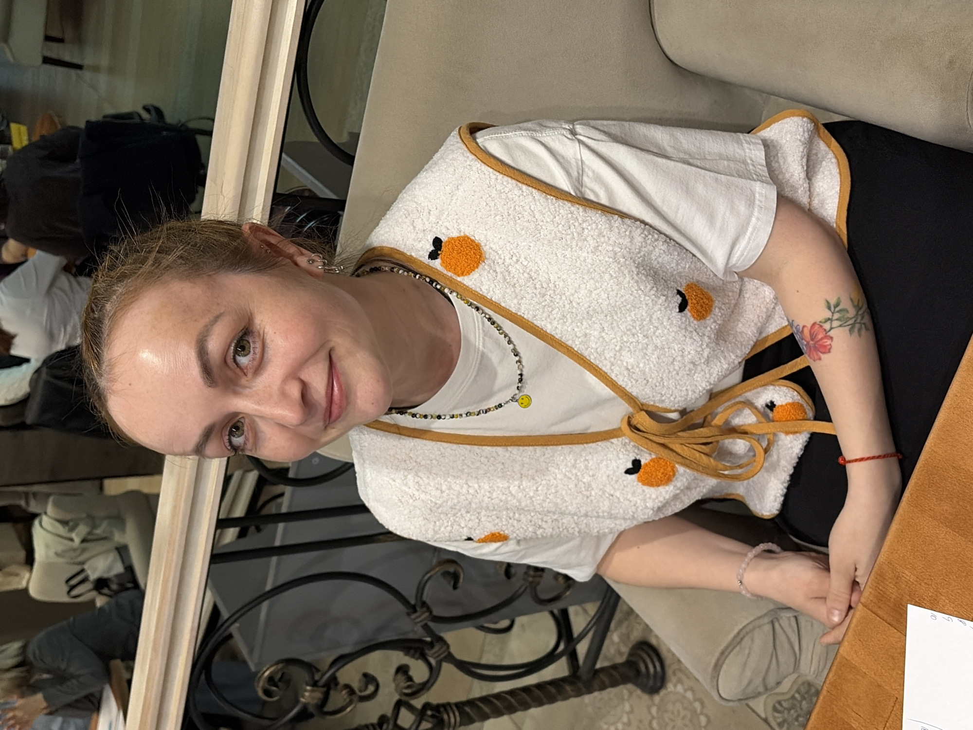 A woman is seated at a table, likely in a cafe or restaurant, with a distinctive white vest decorated with orange-shaped appliques and tied at the front. She is wearing a white t-shirt underneath, and a beaded necklace with a smiley-face charm. Her left arm has a flower tattoo and a red string bracelet. She has fair skin, light brown hair pulled back, and is looking at the camera with a slight smile. The table is made of light-colored wood. The backdrop features dark metal furniture and a mirror reflecting other people. The location is in Chaikovsky, Russia.