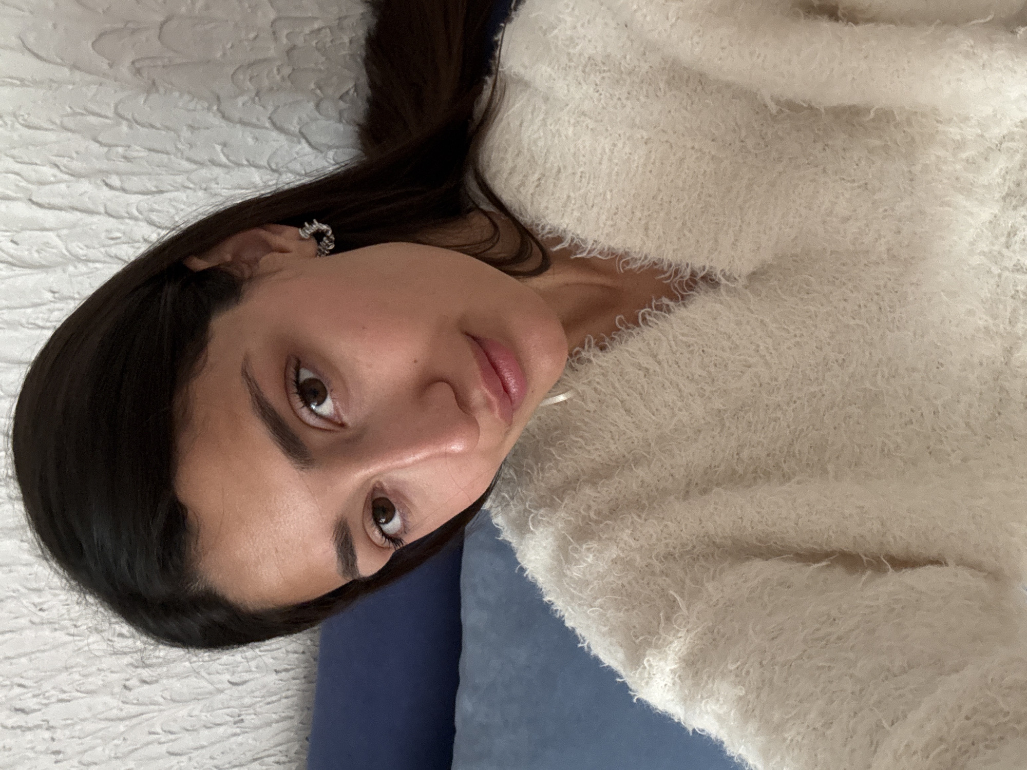 A woman with dark hair and brown eyes is lying on her back. She is wearing a fluffy, off-white sweater. She has a small earring in her left ear. She is lying on a textured white surface with a blue pillow beneath her head. The location is not provided in the image.