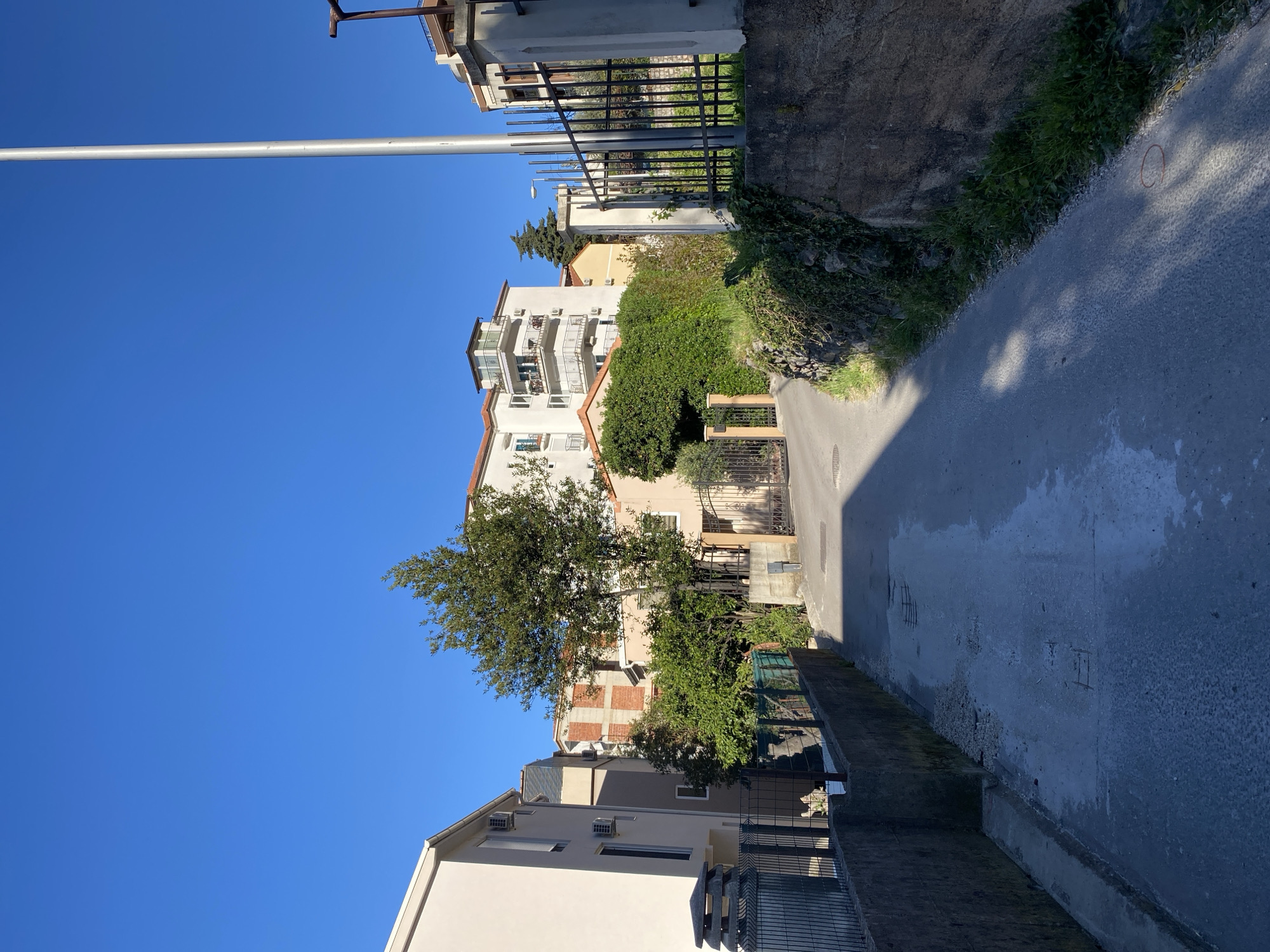 The image shows a street in Budva, Montenegro, lined with residential buildings. The sky is clear and blue. A tall metal pole stands to the left. A narrow paved road stretches into the distance, with walls and fences bordering it. Vegetation is visible along the edges of the road. The architecture of the buildings is typical of the region, with white or beige facades and tiled roofs. Air conditioning units are attached to some of the buildings. There are no people visible in the frame. The overall atmosphere is peaceful and quiet.