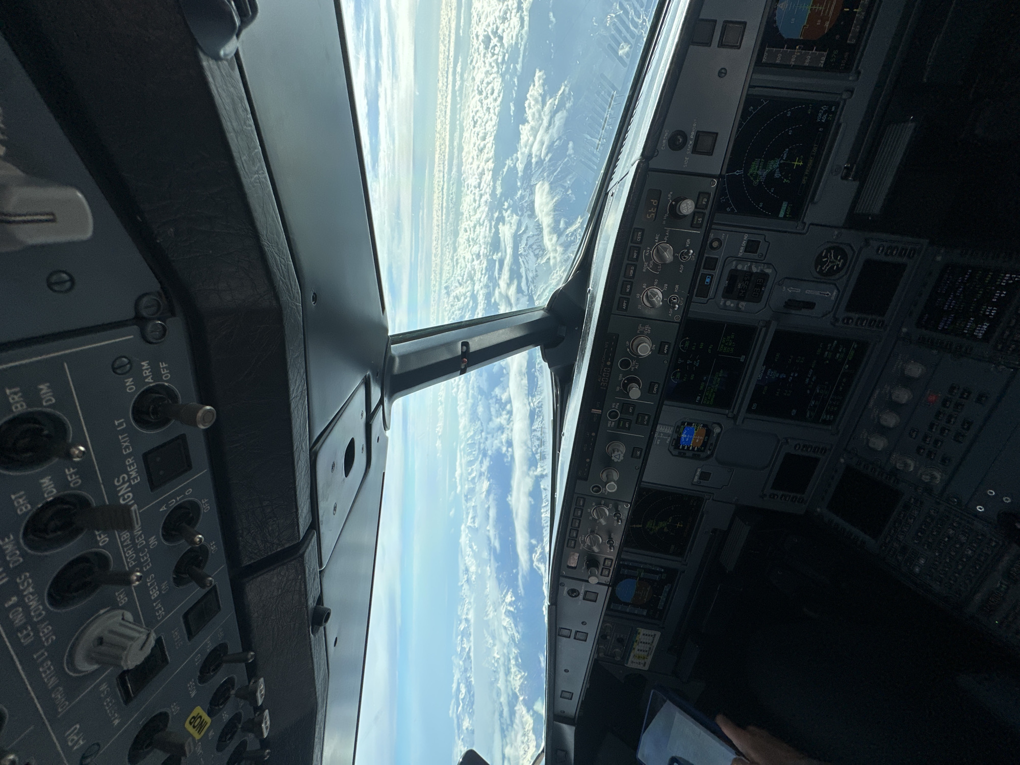 This image shows the interior of an airplane cockpit during flight, as evidenced by the view through the windshield of clouds and terrain below. The cockpit instrumentation includes various control panels, displays, switches, and dials. The control panels are labeled with terms like "EMER EXIT LT," "SEAT BELTS," "MASTER SW," "APU," "NAV," "VOR," "LS," "FD," "CHRONG," "SIDE STICK PRIOR," "TERR ON ND," and "CSTR," all typical of aircraft controls.
There is no visible evidence in the image to suggest the location is Sochi, Russia.