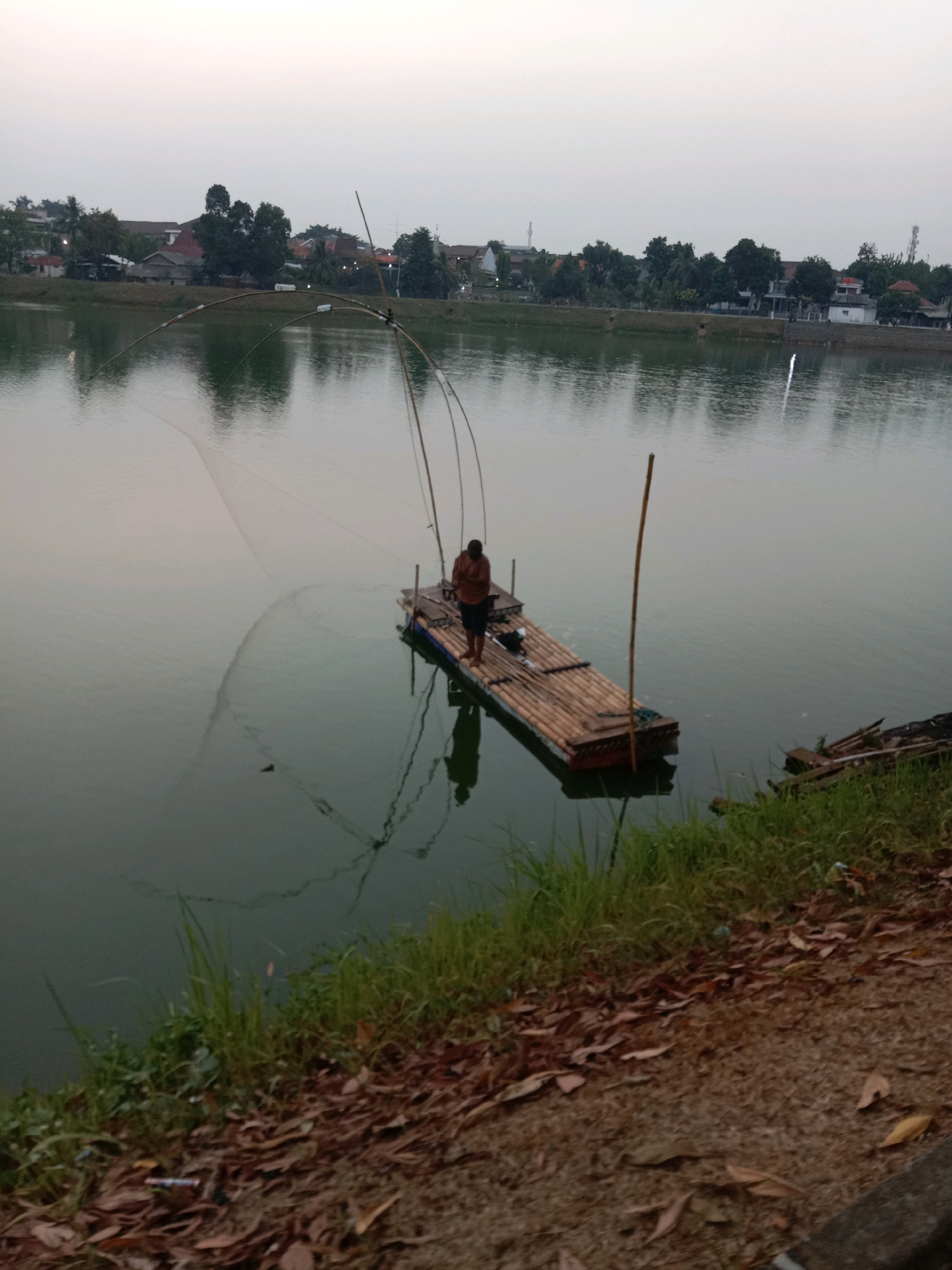 A person stands on a bamboo raft in a lake, holding a fishing net. The raft is equipped with two tall poles, one of which is topped with a fishing net. The lake is calm and reflects the sky and the surrounding buildings. In the background, there are houses and trees along the shore. The scene appears to be in a suburban or semi-rural area, likely in South Tangerang, Indonesia, given the architectural style of the buildings and the presence of tropical vegetation. The sky is overcast, suggesting it is either early morning or late evening. The ground in the foreground is covered with dry leaves and grass.