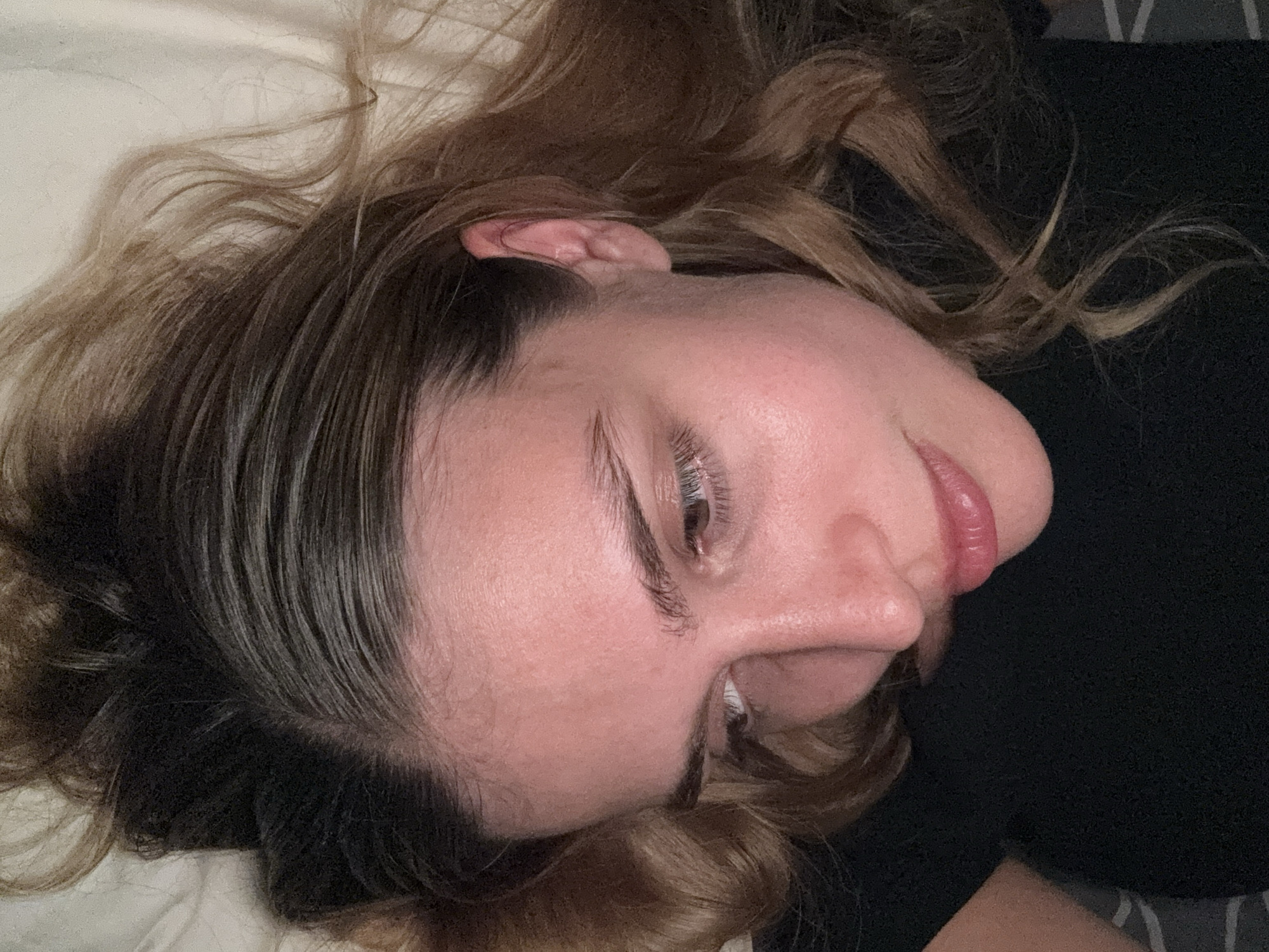 A person lies with their head on a white pillow. The person has light skin and brown hair. The person is wearing a dark-colored shirt. The location is Unknown, Unknown.