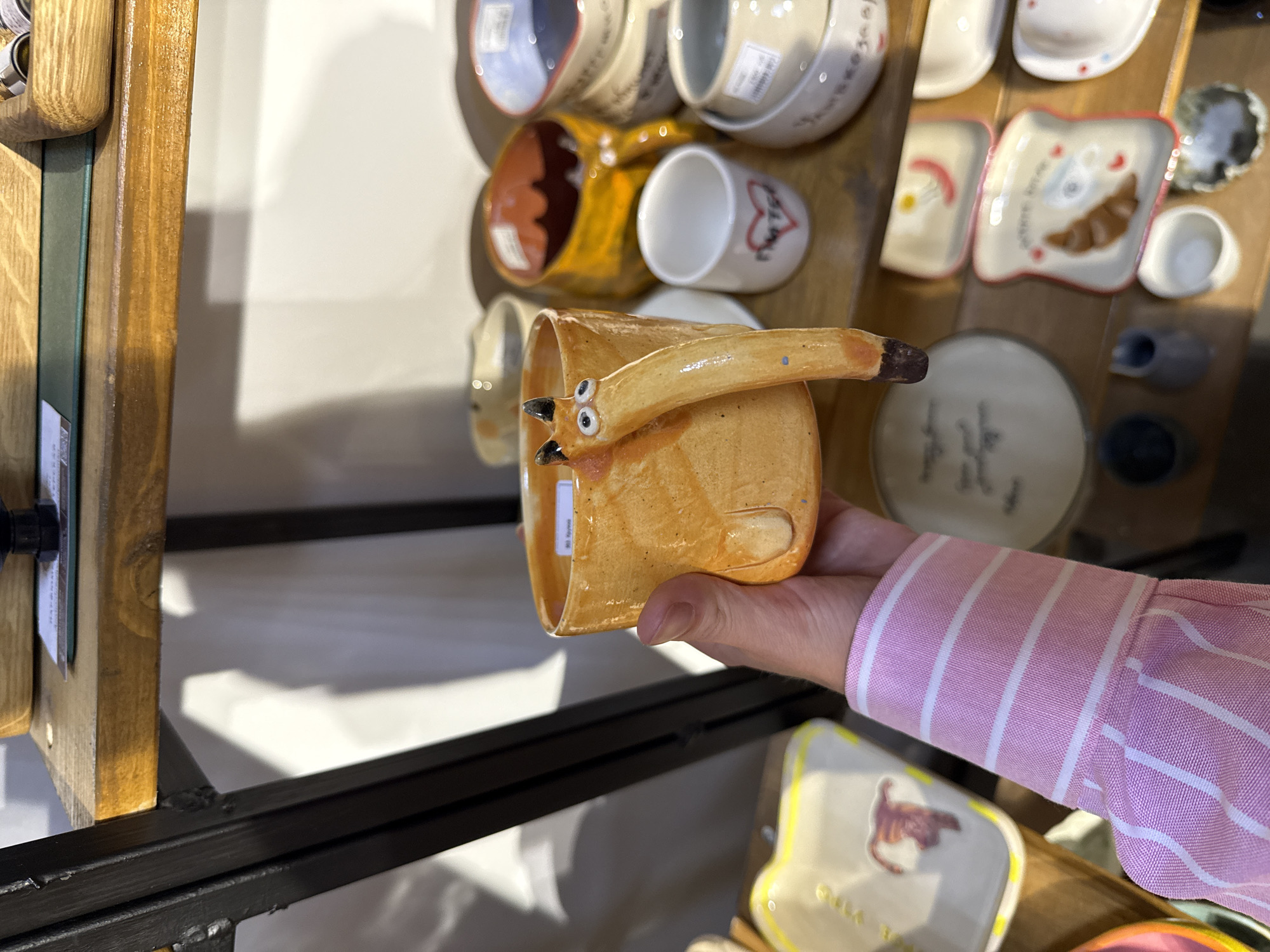 The image shows a person holding a handcrafted ceramic mug with a fox-like handle and eyes. The mug is orange with a textured surface. The person is wearing a pink and white striped shirt. The mug is being held in front of a display of various ceramic items on wooden shelves. These items include plates, bowls, and smaller decorative pieces. The background appears to be a shop or market stall. The lighting is bright and appears to be natural. The scene is likely in Saint Petersburg, Russia, given the context of the request.