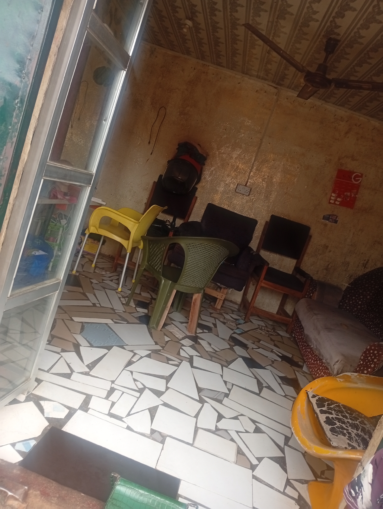 The image captures an indoor scene, likely a reception area or waiting room, in Jalingo, Nigeria. The room has a rustic feel with rough, textured walls painted in a yellowish-brown hue. The ceiling is covered with a patterned wallpaper, and a brown ceiling fan with three blades is visible. The floor is tiled with irregularly shaped white, beige, and light blue pieces, giving it a mosaic-like appearance.
Several chairs are arranged in the room. In the foreground, to the right, is a bright yellow plastic chair with a patterned cushion inside. To its left, a green plastic chair is positioned. Further back, there are two dark-colored chairs with dark upholstery and wooden frames. Another yellow plastic chair with metal legs is visible in the middle ground.
On a shelf behind a glass door on the left side of the image, various items are stored, including what appears to be bags and containers. A red poster with white text and a "G" logo is affixed to the wall on the right. An electrical outlet is also visible on the wall. A portion of a sofa with a patterned fabric is seen on the far right of the frame, partially obscured by the yellow chair. The overall impression is of a functional, perhaps slightly cluttered, waiting area.