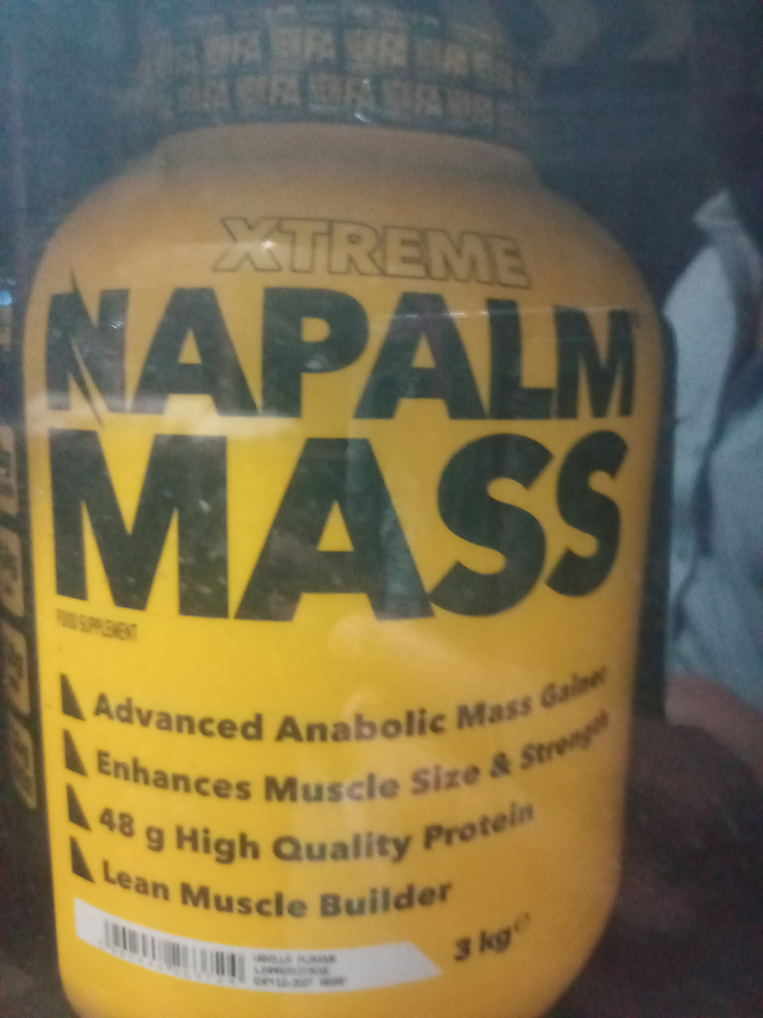 A yellow cylindrical container, branded "XTREME NAPALM MASS," is depicted in a close-up, slightly angled shot. The product is identified as a "FOOD SUPPLEMENT" and features four key attributes listed in black text: "Advanced Anabolic Mass Gainer," "Enhances Muscle Size & Strength," "48 g High Quality Protein," and "Lean Muscle Builder." The net weight, "3 kg," is visible at the bottom right of the label. A white label at the base of the container displays a barcode and small print, including "VANILLA FLAVOR" and partially legible alphanumeric characters, one of which appears to be an expiration date or batch number. The background is dark and out of focus, suggesting an indoor setting, with faint, repetitive text patterns visible at the top behind the container, possibly from other packaging or shelving. No people are visible.