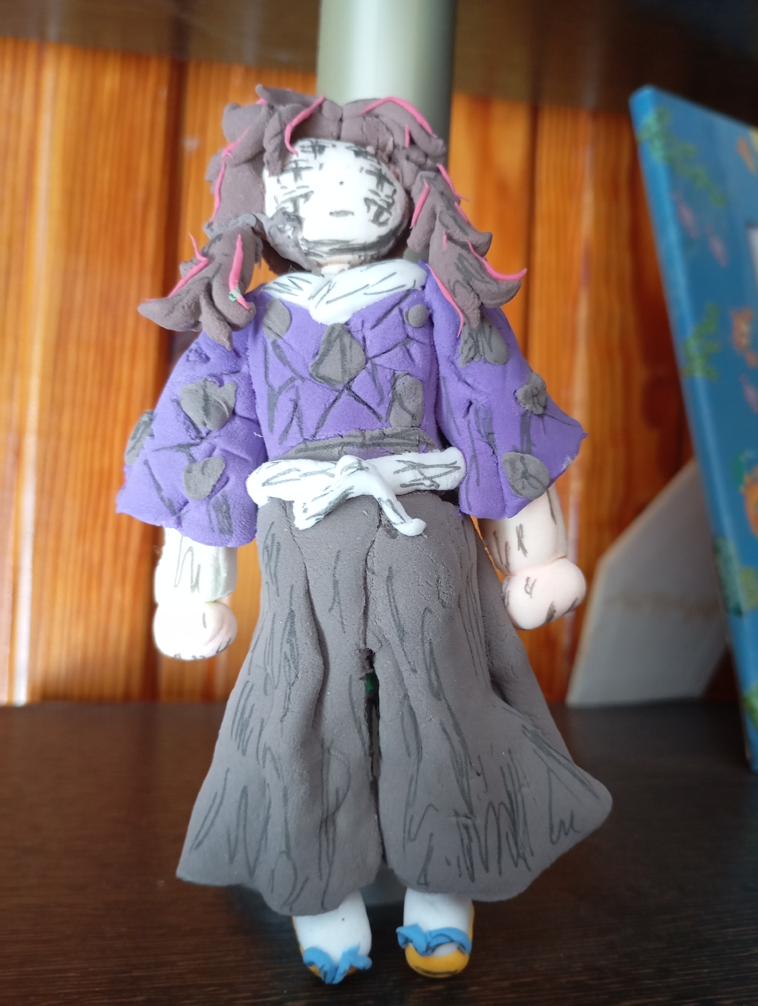 A clay figure of a person with dark, messy hair and pink highlights stands on a dark wooden surface. The figure wears a purple jacket with grey leaf-like embellishments and a grey skirt. The face of the figure is white with black markings. The background consists of a wooden plank wall. A blue object with green designs is visible to the right of the figure. The location is the city of Льниха, Russia.