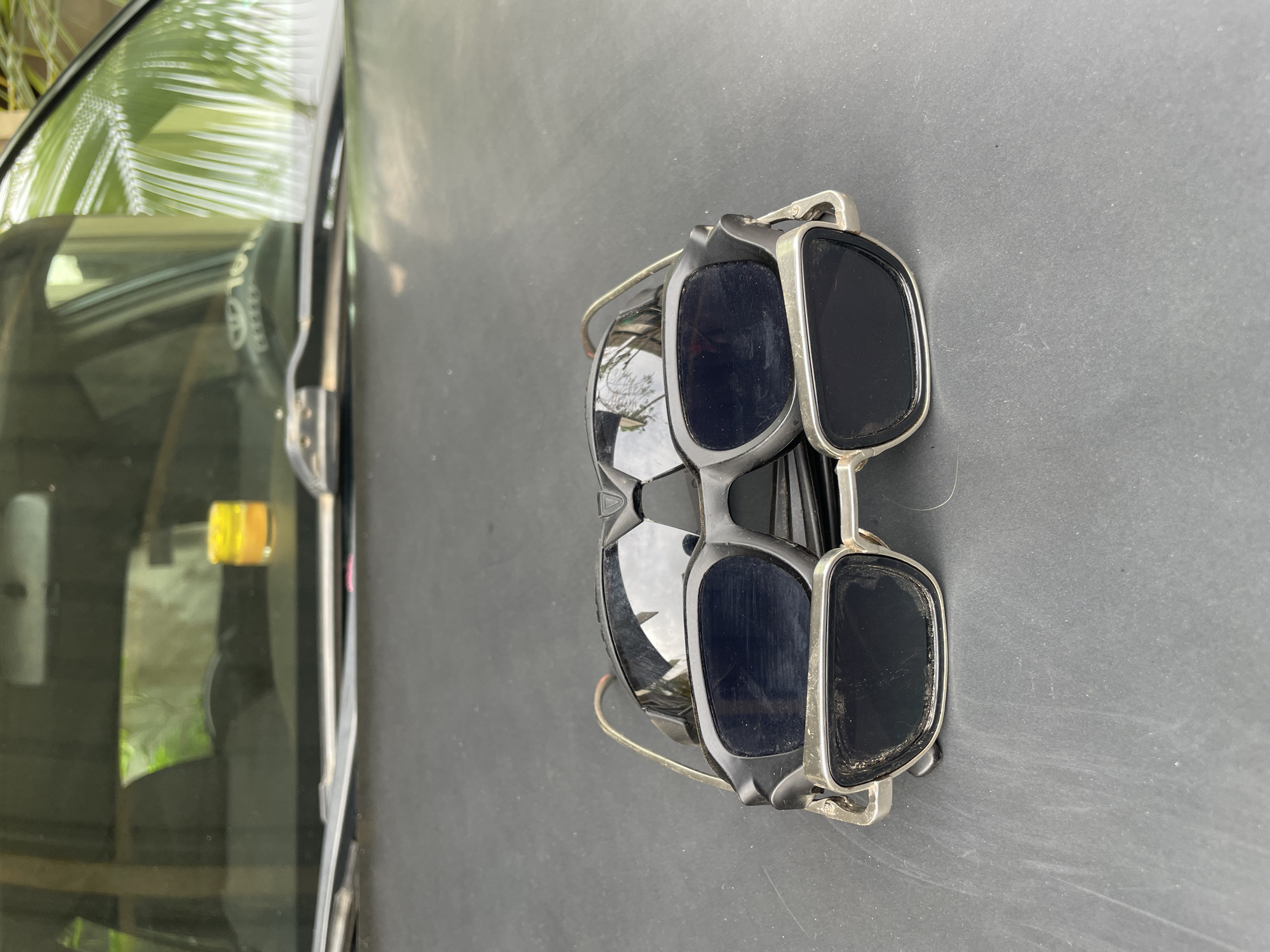 Two pairs of sunglasses are stacked on a dark, possibly black, surface. The pair on top has rectangular lenses and a silver frame. The pair underneath has rounded lenses and a dark frame. The sunglasses are positioned on the hood of a vehicle, as evidenced by the reflection of a tree and a yellow object on the car's windshield. The car appears to be a Toyota, as suggested by the logo visible on the windshield. Given the prompt, the location is Akwanga, Nigeria.