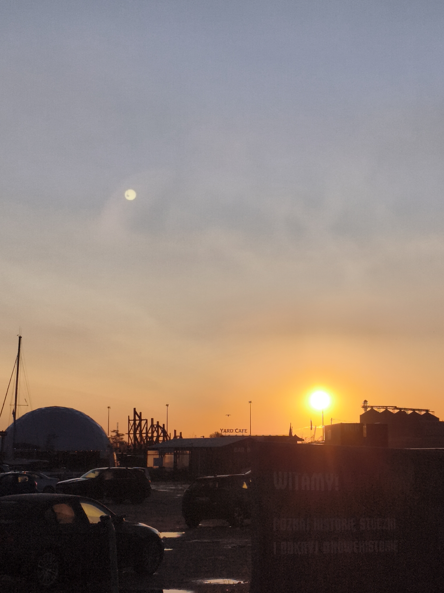 The image depicts a harbor area in Gdańsk, Poland, at sunset. The sky transitions from a pale blue at the top to a vibrant orange and yellow near the horizon. A faint, pale moon is visible in the upper left quadrant of the sky. Silhouetted against the sunset are several structures including what appears to be a domed building, a tall mast of a sailboat, industrial silos, and various buildings that might be part of the port or associated businesses, one of which has a sign reading "YARD CAFE". In the foreground, several dark-colored cars are parked on what appears to be a wet surface, possibly due to recent rain, with puddles reflecting the twilight. There are no people clearly visible in the image. The overall scene suggests a maritime or industrial port setting during the late evening.