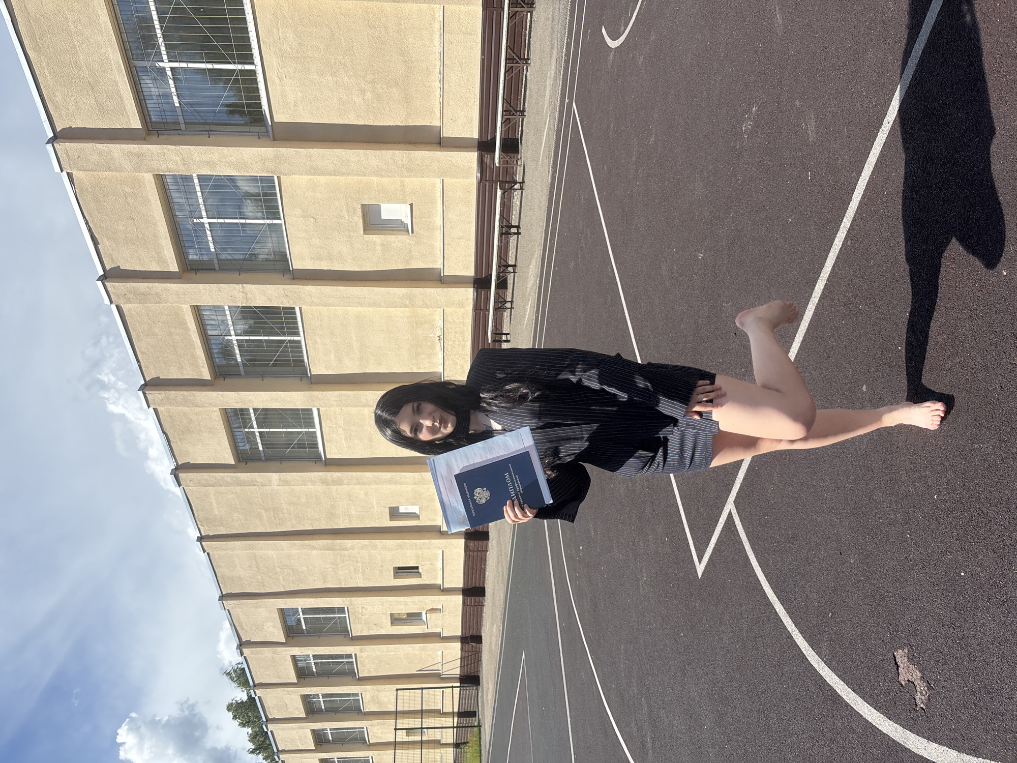 A young woman, appearing to be of Russian ethnicity, stands on an asphalt sports court, possibly a basketball court, given the painted lines. She is wearing a dark pinstripe suit jacket and skirt. Her right leg is bent at the knee, and her left leg is extended. She is barefoot. She holds a blue diploma in her hands, which are positioned in front of her. The diploma has visible Russian text.
Behind her is a light-yellow, multi-story building with numerous windows, many of which have metal security bars. The building's architecture suggests it might be a school or public institution. The time of day appears to be daytime, with a bright sky visible, indicating sunny weather with some clouds. The location is Chaikovsky, Russia. The woman's expression is neutral or slightly serious.
