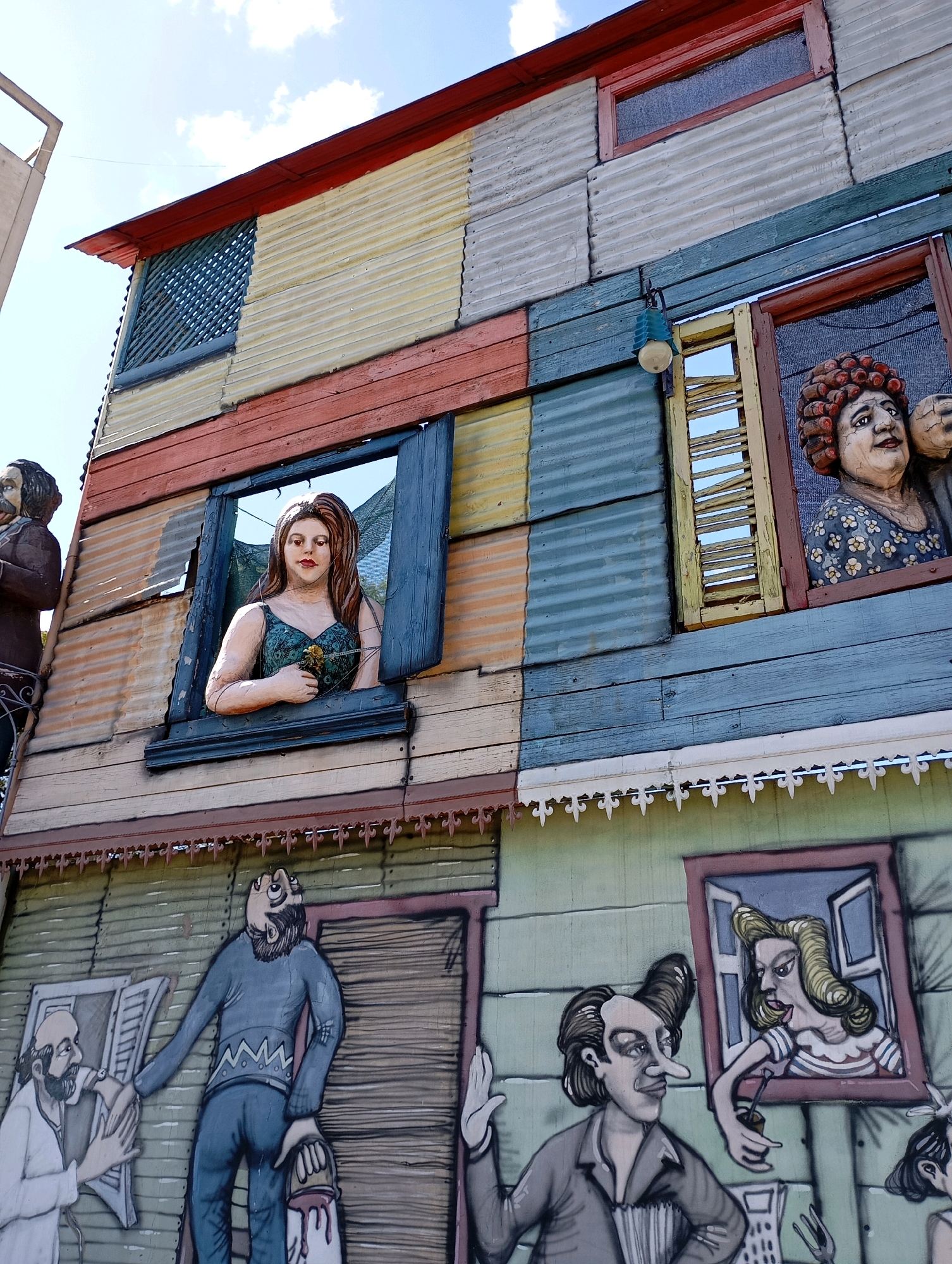 This image depicts the colorful and vibrant architecture of Caminito, a pedestrian street and museum in Buenos Aires, Argentina. The buildings are constructed from corrugated metal and brightly painted wood, creating a patchwork effect. Several windows feature three-dimensional statues of people looking out, adding a unique theatrical element.
On the upper level, a statue of a woman with long brown hair and a green dress is visible in a window. To her right, another statue of a woman with curly red hair and a floral dress is perched in a window. Both statues appear to be holding or interacting with unseen objects.
The lower level features a mural depicting several figures. On the left, a man with a beard, possibly a doctor, is shown examining or interacting with another man dressed in blue. This man is holding a paint bucket with dripping paint. To the right of this mural, another mural shows a man with distinctive styled hair and a prominent nose, playing an accordion. He is looking towards a window where a woman with blonde hair is leaning out, holding a straw.
The sky above is a clear blue with scattered white clouds. The overall scene conveys a lively and artistic atmosphere, characteristic of this famous Buenos Aires landmark.