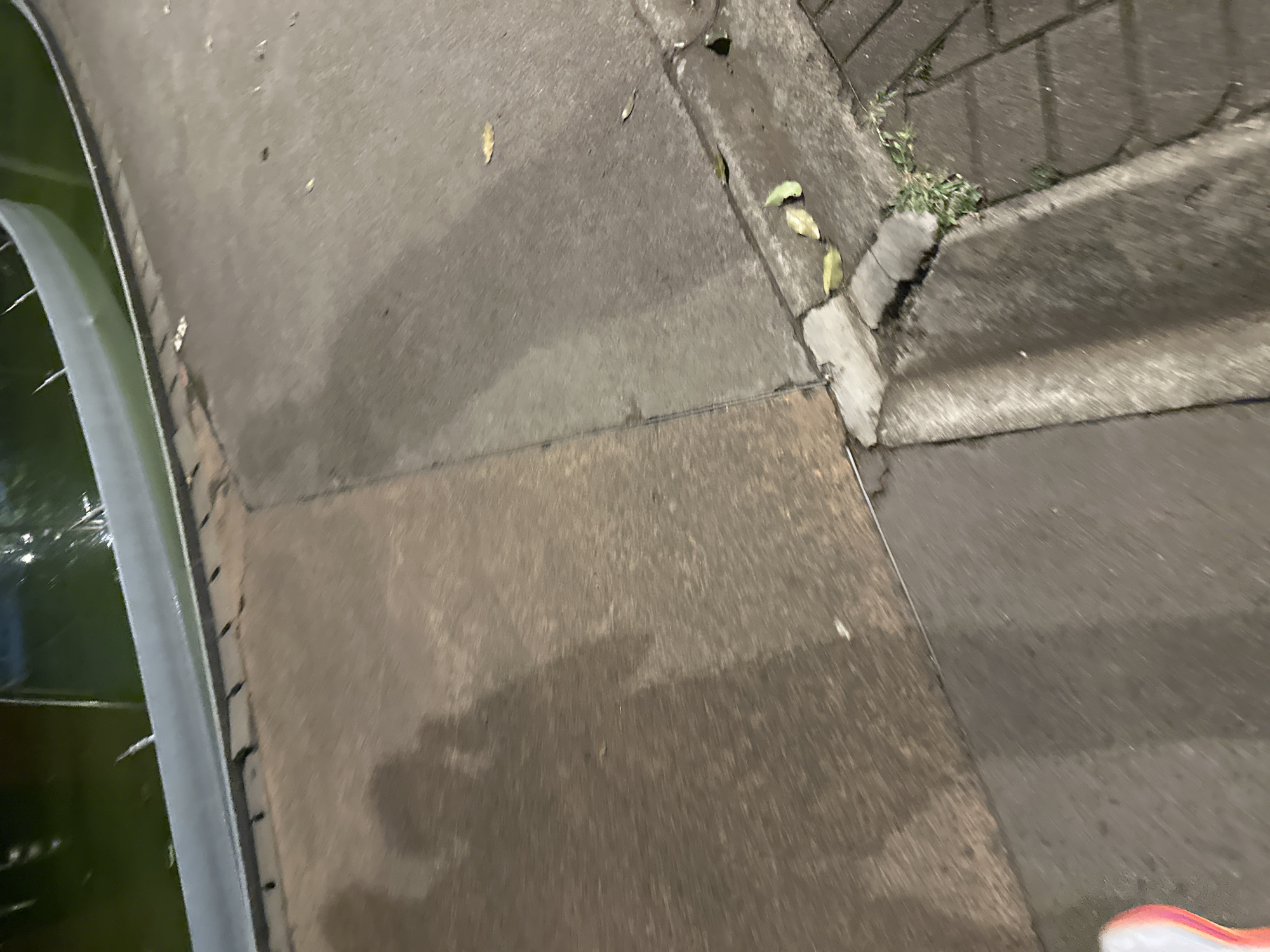 The image depicts an outdoor urban scene. A sidewalk with concrete segments leads up to a set of steps constructed from similar material. The sidewalk surface appears damp in areas. Along the left edge, there's a structure with visible metal elements and possibly reflective material, indicating a body of water or shiny surface. Some plants grow near the edge of the steps. A glimpse of a shoe is visible in the bottom right corner. The location is indicated as Central Jakarta, Indonesia. No people or overt actions are observed in the frame.