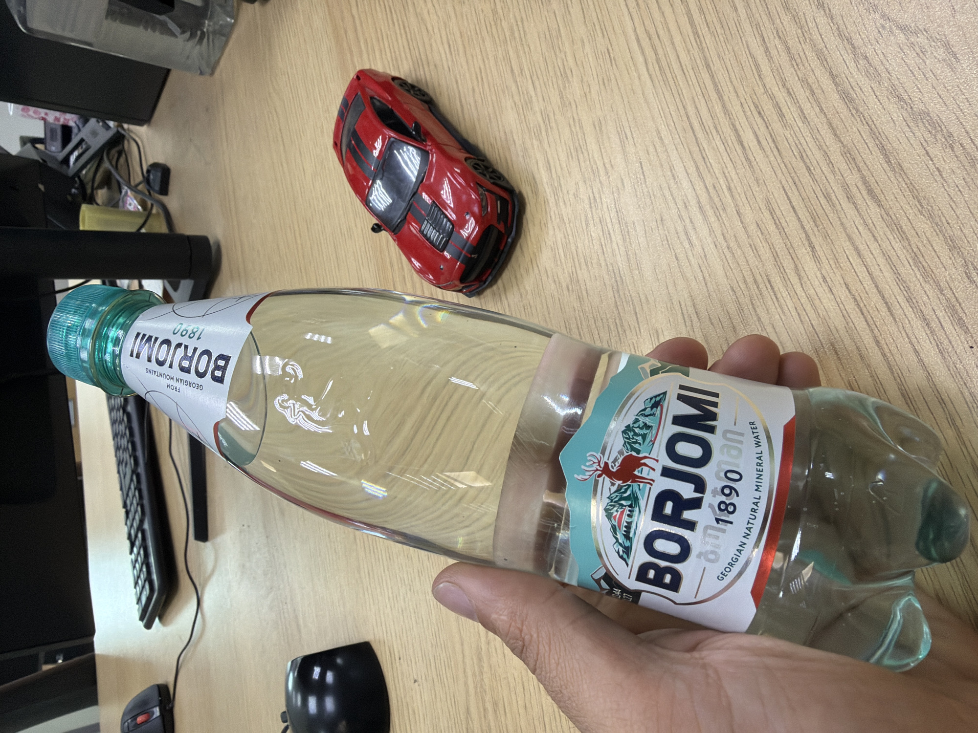 A hand holds a clear plastic bottle of Borjomi mineral water on a wooden desk. In the background, a red toy car sits near a computer monitor and keyboard. A computer mouse and a second, unidentified black object are also visible on the desk. The location of the scene is not discernable from this image.