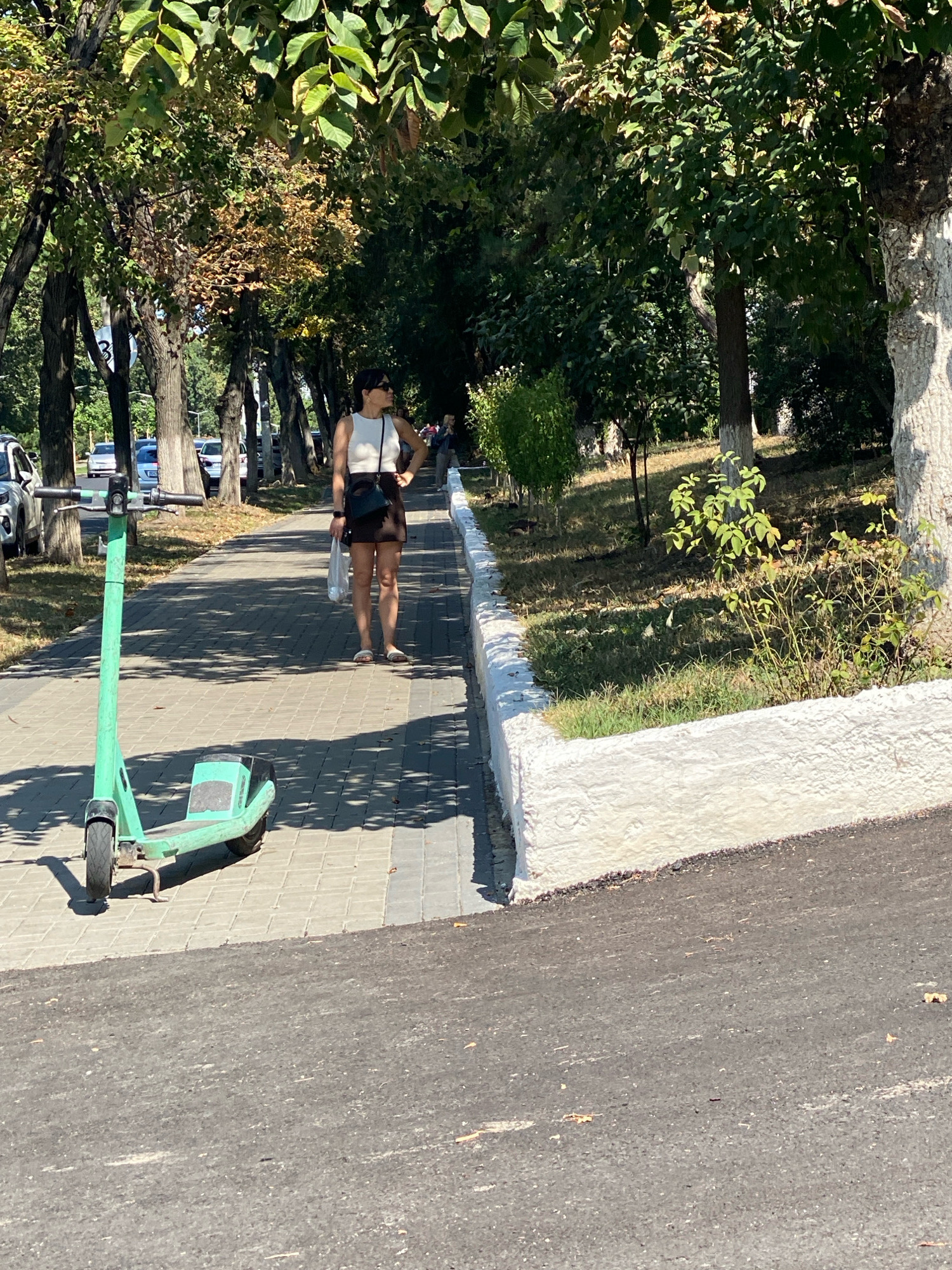 The image depicts a sun-drenched sidewalk lined with trees in Chișinău, Moldova. A woman, dressed in a white crop top, dark skirt, and sandals, stands on the paved walkway, holding a white plastic bag. Her right hand is on her hip, and she appears to be looking to her left. In the foreground, a green electric scooter is parked on the sidewalk. The walkway is bordered by a white-painted curb on the right, beyond which is a grassy area with some bushes. Cars are visible parked along the street to the left of the sidewalk. The overall scene suggests a typical summer day in a city park or along a tree-lined avenue.
