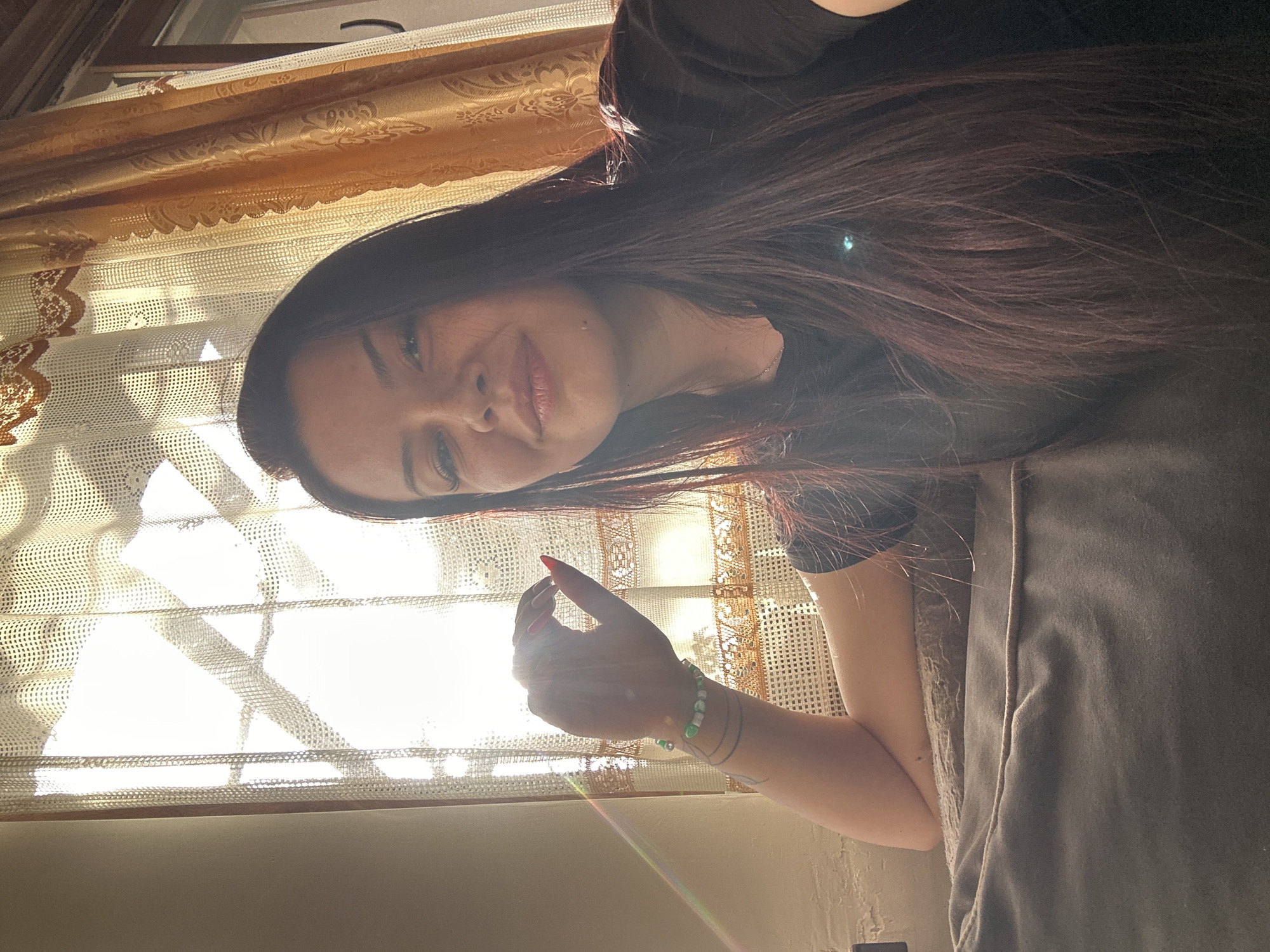 A woman with long, dark hair is shown in a close-up, angled shot. She is wearing a dark-colored t-shirt and has a green beaded bracelet on her wrist. She is positioned near a window covered with light-colored, patterned curtains. The window appears to be letting in sunlight. The image does not contain information to determine if the photo was taken in the city of Skala, Bulgaria.