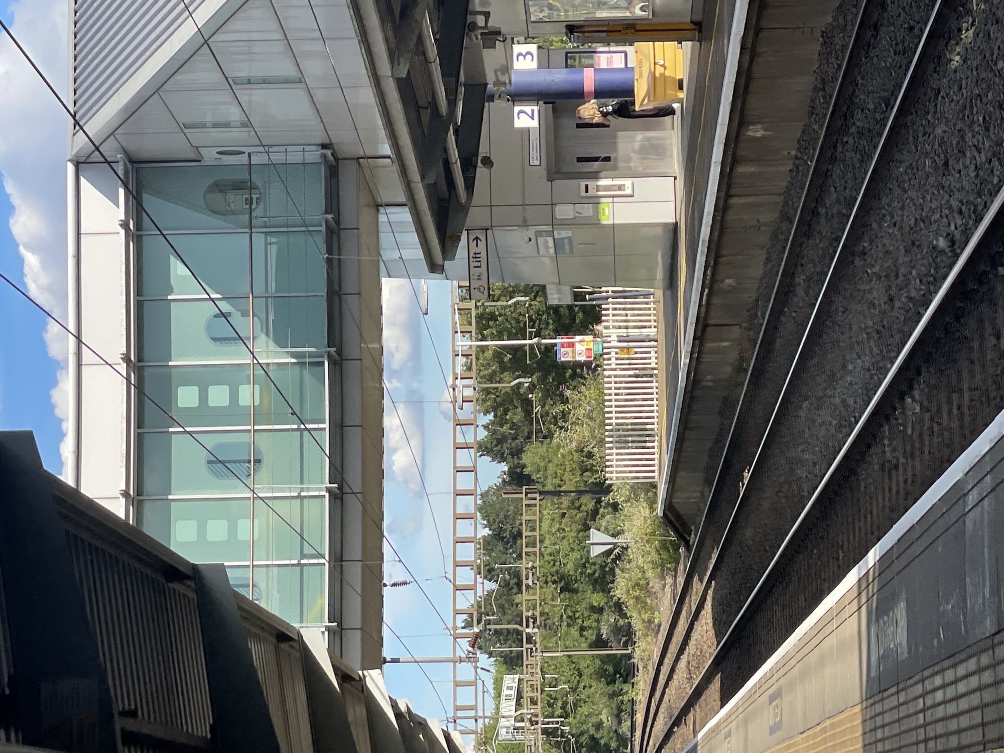 This is an eye-level, medium shot of a train station in Luton, United Kingdom. The scene is oriented vertically, with the tracks running diagonally from the foreground to the background, suggesting a slight downward angle. Sunlight streams down, indicating it is daytime with clear skies and fair weather.
The foreground features a train platform with dark gray, textured surfaces and yellow markings. Next to it, parallel train tracks are visible, filled with dark gravel and gleaming steel rails. To the left, a modern building with a glass facade and horizontal louvers dominates the left side of the frame. The building's reflective windows offer glimpses of interior structures.
In the middle ground, a walkway or bridge extends across the tracks, connecting to the building. A woman with blonde hair is partially visible near a lift entrance, indicated by a sign with a directional arrow and the word "Lift." Above the lift, numbers "2" and "3" are displayed, possibly indicating platform or level numbers. Other signs with text are present but difficult to decipher.
The background is filled with lush green trees, suggesting a natural element within the urban environment. Overhead, electrical wires and support structures for the train line are visible against a bright blue sky with scattered white clouds. The overall impression is one of a functional, modern transportation hub set against a backdrop of nature on a sunny day.