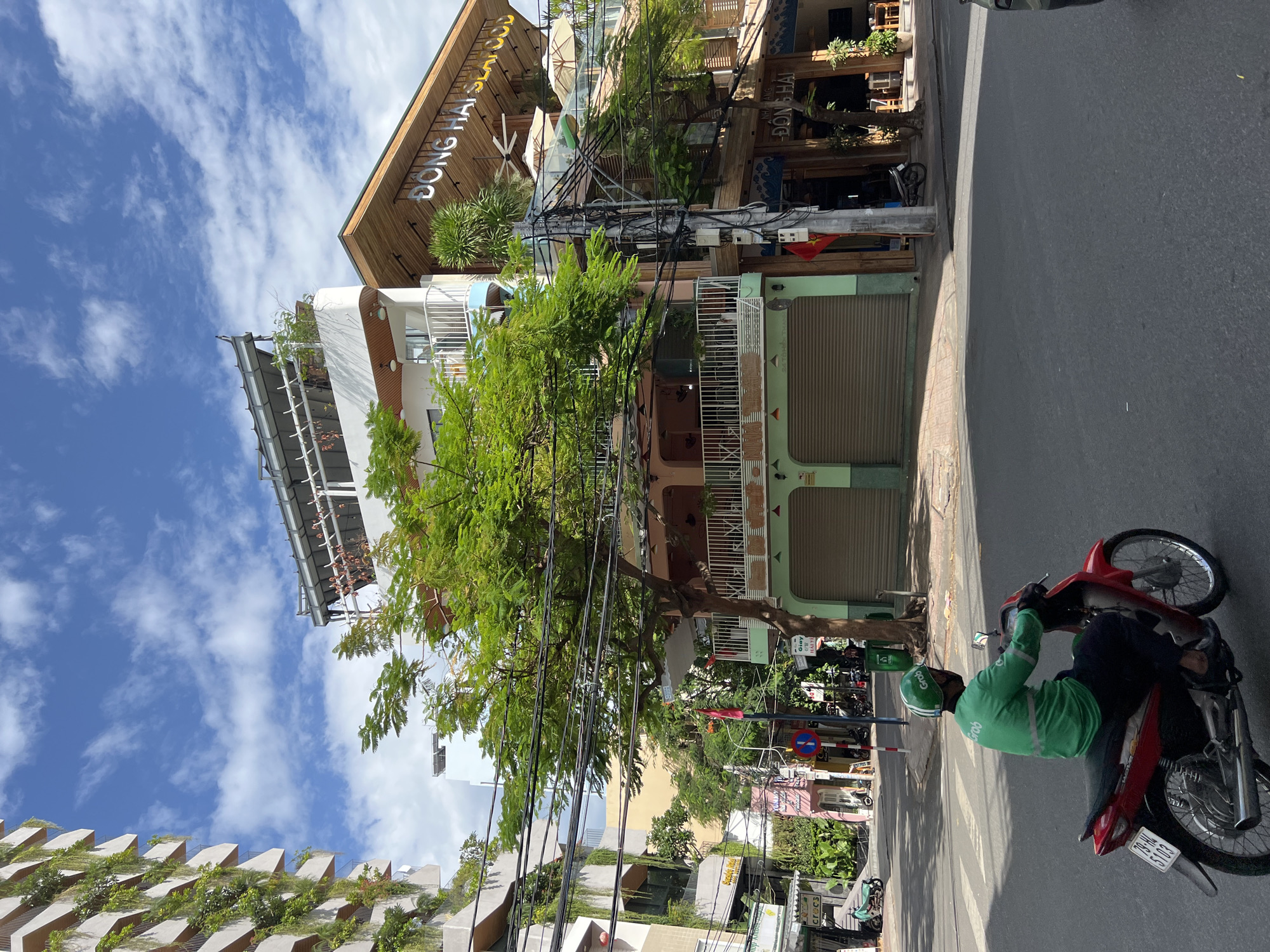 A Grab delivery driver on a red motorcycle is turning at an intersection in Nha Trang, Vietnam. The driver is wearing a green Grab uniform and helmet, and a black face mask. The motorcycle has a white license plate with black lettering. In the background, there are several buildings, including a modern building with plants on its exterior, and a restaurant with a sign that reads "DONG HAI SEAFOOD". The sky is blue with white clouds. The street is paved with asphalt.