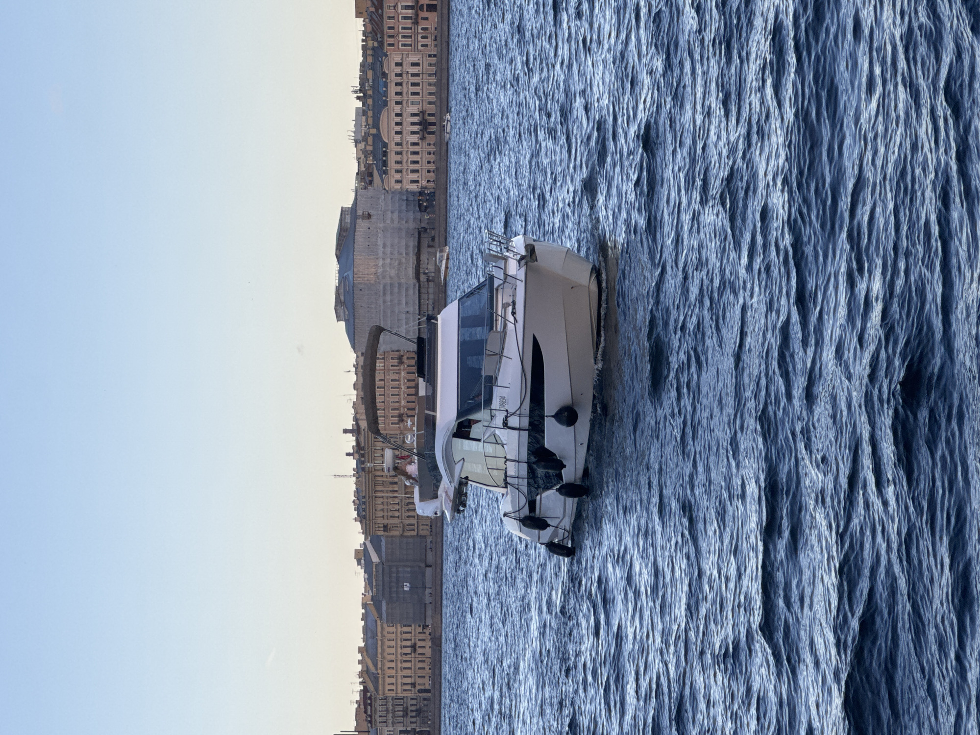 A white motor yacht is navigating on the Neva River in Saint Petersburg, Russia. The yacht has a covered seating area and a small white outboard motor. Several black fenders are attached to its port side. In the background, a row of historic buildings line the riverbank. The sky is clear and a pale blue. The water is a deep blue with moderate waves. There is a person in the yacht's cockpit.