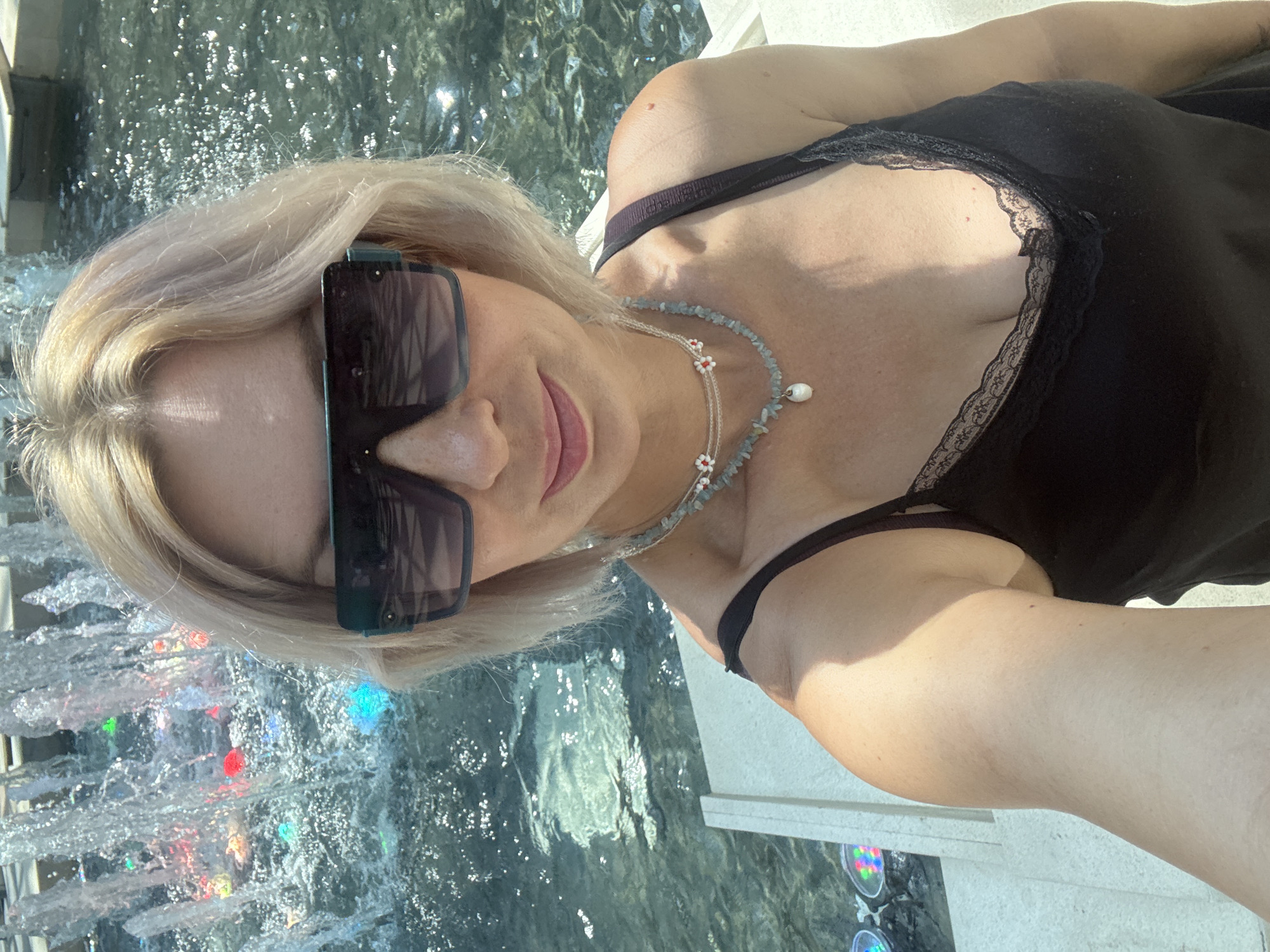 A woman with short, blonde hair and sunglasses is taking a selfie in front of a fountain in Sochi, Russia. She is wearing a black tank top with lace trim and two necklaces. The fountain has colorful lights. The water is reflecting the sunlight.