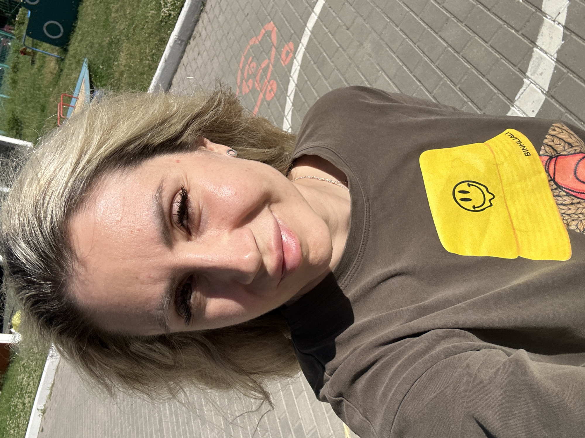 A medium shot captures a woman with blonde hair styled loosely. She is looking at the camera and smiling, with sunlight casting shadows on her face. She is wearing a dark brown t-shirt with a yellow bucket hat graphic featuring a smiley face. She is also wearing a silver necklace. The background shows a paved area with a white line and an orange symbol that appears to be an electric vehicle charging station marker. Beyond the paved area is a grassy patch with some small white flowers and playground equipment, including a green structure with a circular cutout and a red and blue play structure. The scene is outdoors, suggesting a public space like a park or playground. The overall lighting indicates it is daytime. The location is specified as Novoaltaysk, Russia.