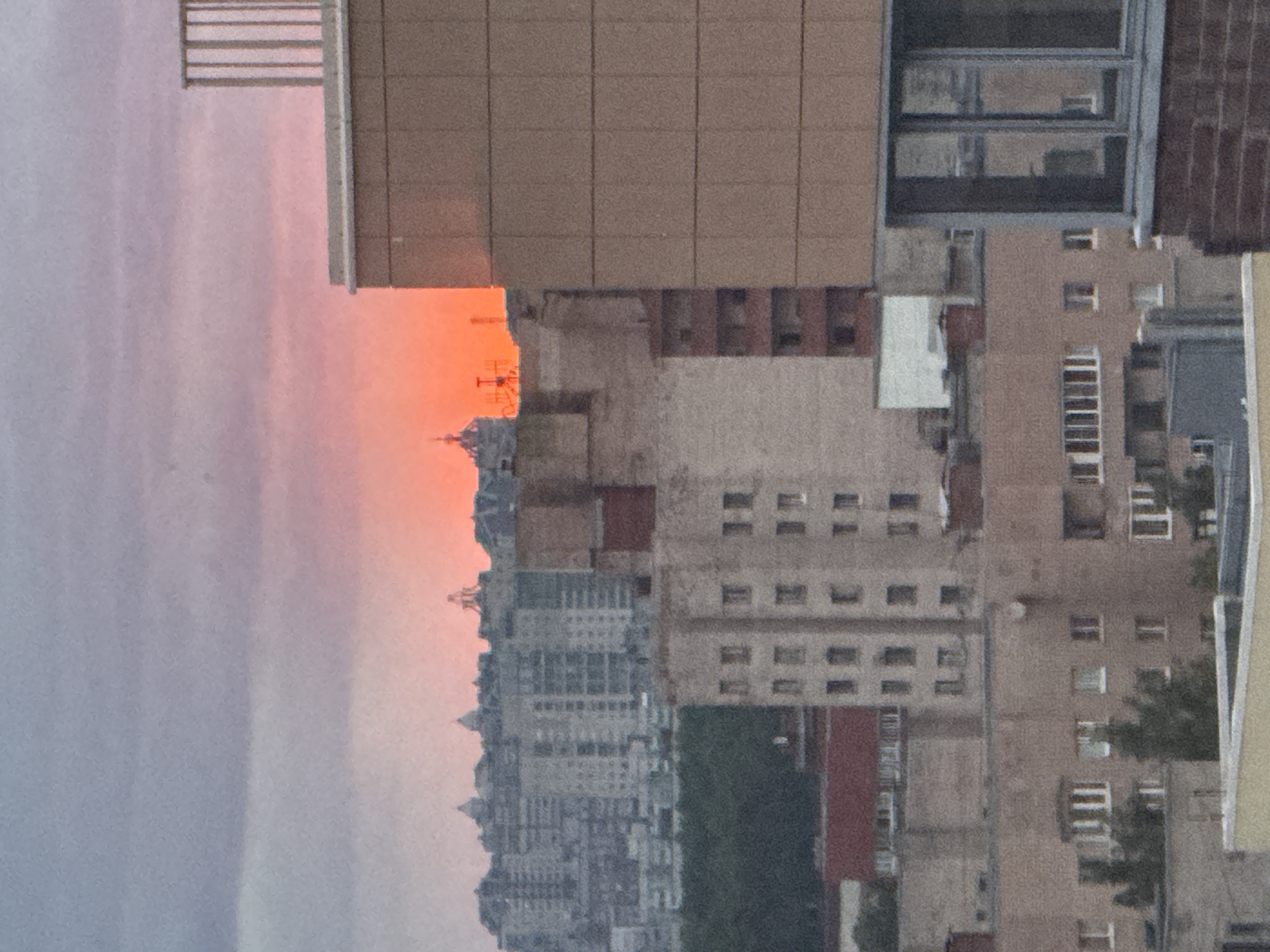 The image shows a cityscape at sunset. In the foreground are parts of buildings constructed with a mix of materials including brick, concrete, and glass. An orange hue, likely from the setting sun, fills the gap between buildings in the distance, and illuminates a communication tower on a far off roof. The background is filled with dense urban buildings and a faintly pink sky. It is impossible to pinpoint the location in Sinyavino, Russia, from the image alone.