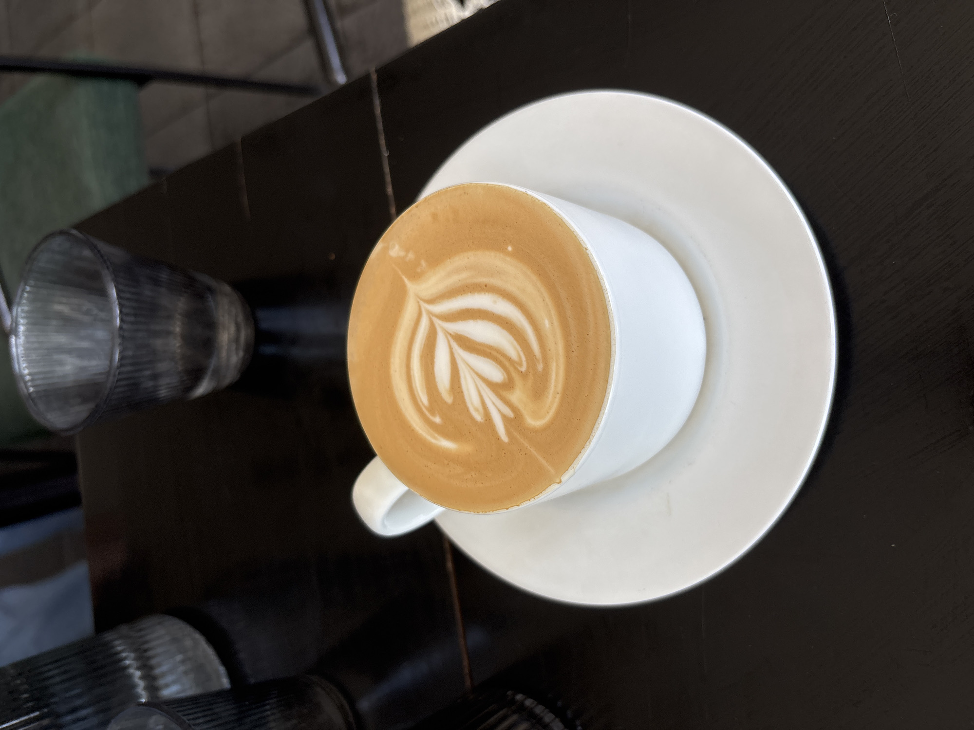 A high-angle shot presents a freshly prepared latte in a white ceramic cup, centered on a matching white saucer, set against a dark table. The latte features intricate, well-defined white foam art, creating a pattern resembling a rosetta or fern, contrasting with the light brown coffee. To the upper left of the cup, a ribbed, clear glass is partially visible on the dark surface. The table itself is dark, possibly wood or laminate, reflecting some light. The background is softly blurred, showing hints of green and darker tones, suggesting an indoor cafe or restaurant environment in Kuta Selatan, Indonesia. The scene depicts a moment of readiness for enjoying a specialty coffee. No people or visible text are present in the image.